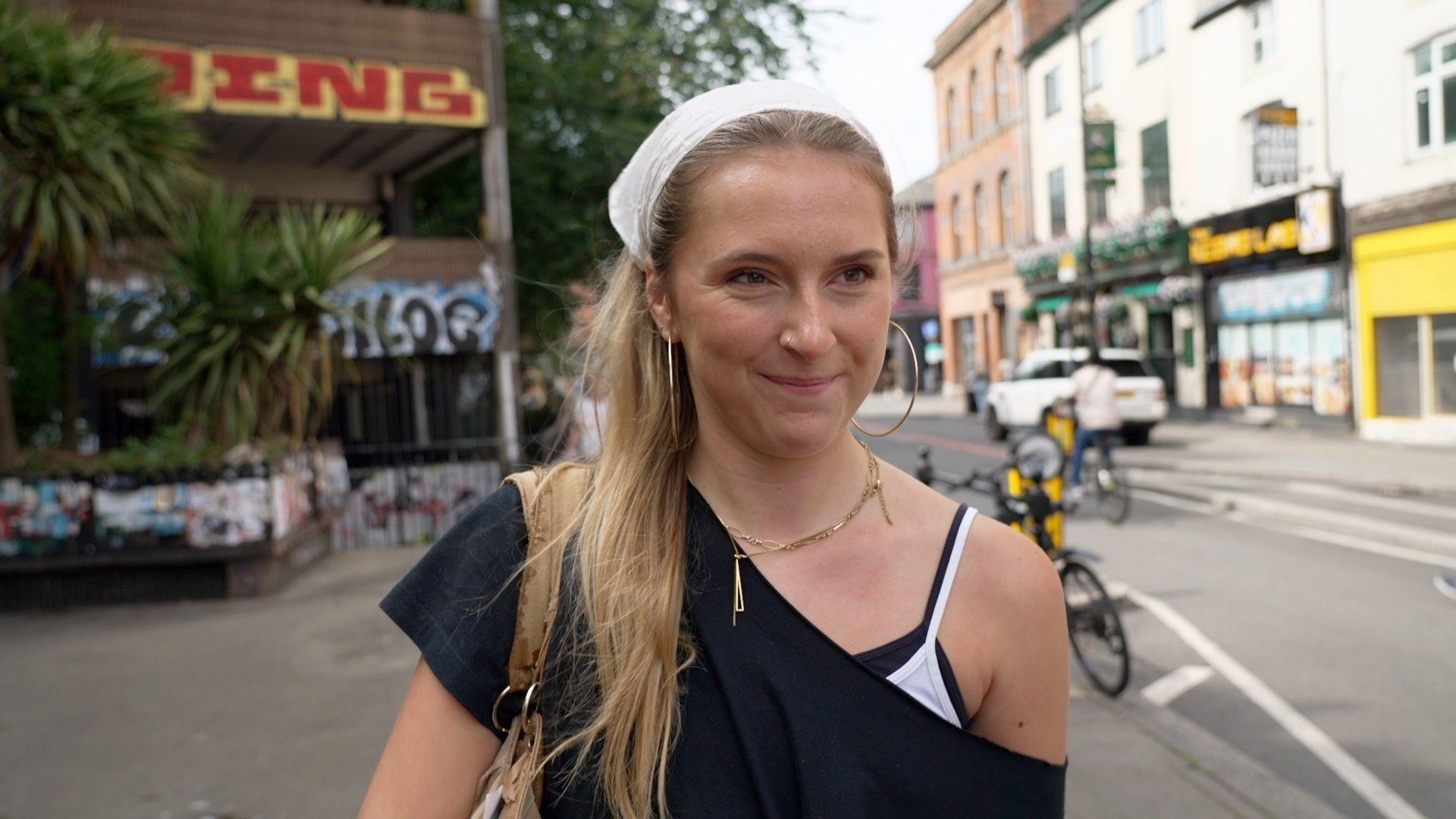 A white woman with long blond hair tied back by a white bandana. She has big hoop earrings and wears a black off the shoulder top and gold necklaces.