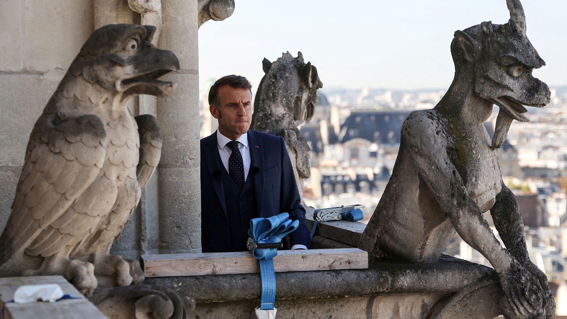 French president Emmanuel Macron, wearing a dark suit stands next to two gargoyle statues on a balcony.