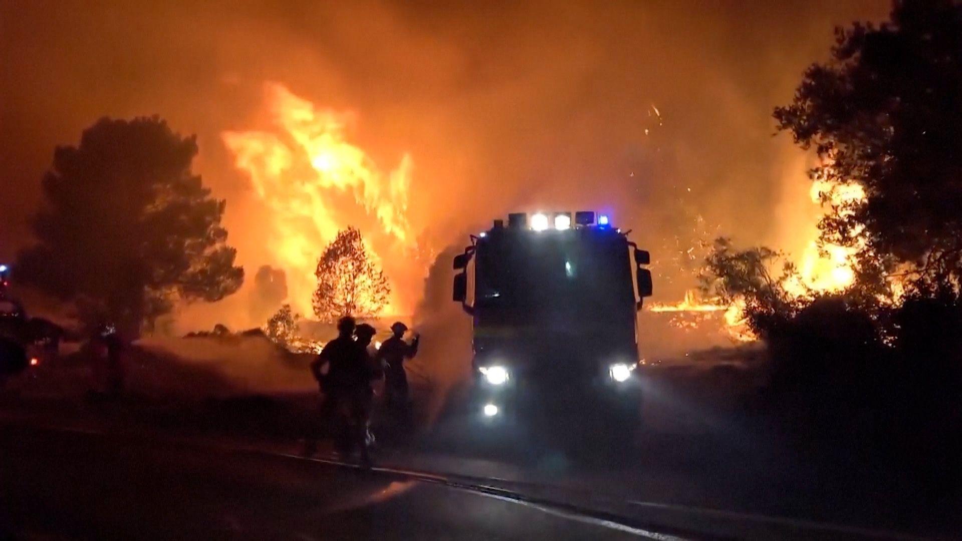 A wall of flames behind a firefighting vehicle and firefighters. It is night and the sky is orange.