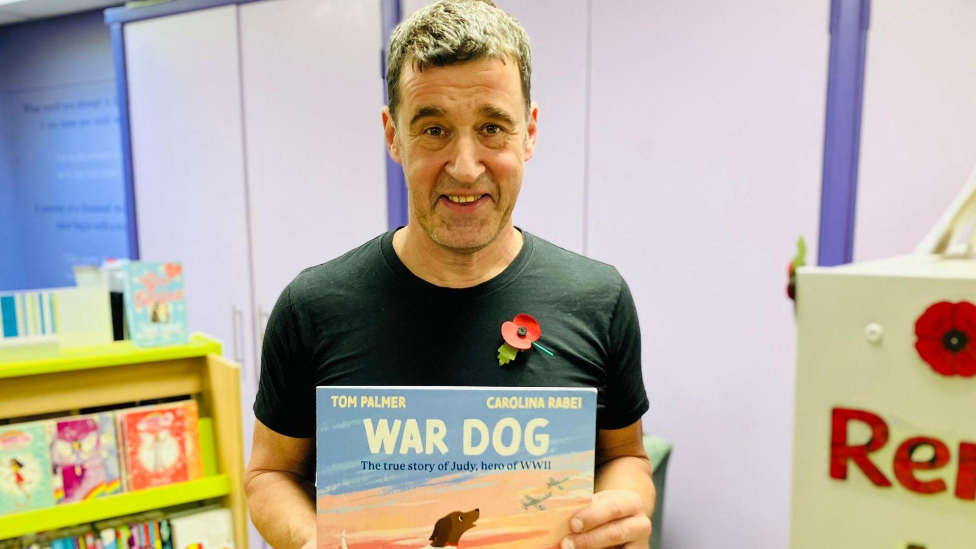 Tom Palmer is wearing a black t-shirt with a red poppy pin. He is standing in a brightly lit library or reading area. He is holding a children’s book titled “War Dog: The true story of Judy, hero of WWII”. The book cover features an illustration of a dog on a boat with a sunset in the background. 
