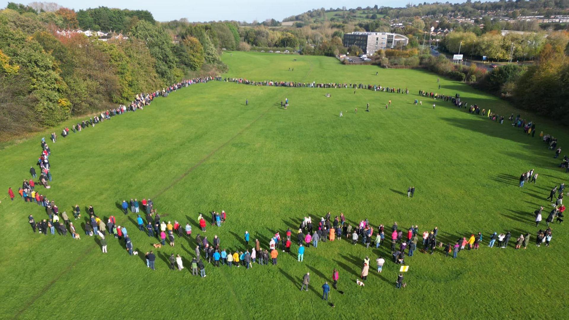 A large circle of demonstrators on a grass park surrounded by trees, as seen from the air