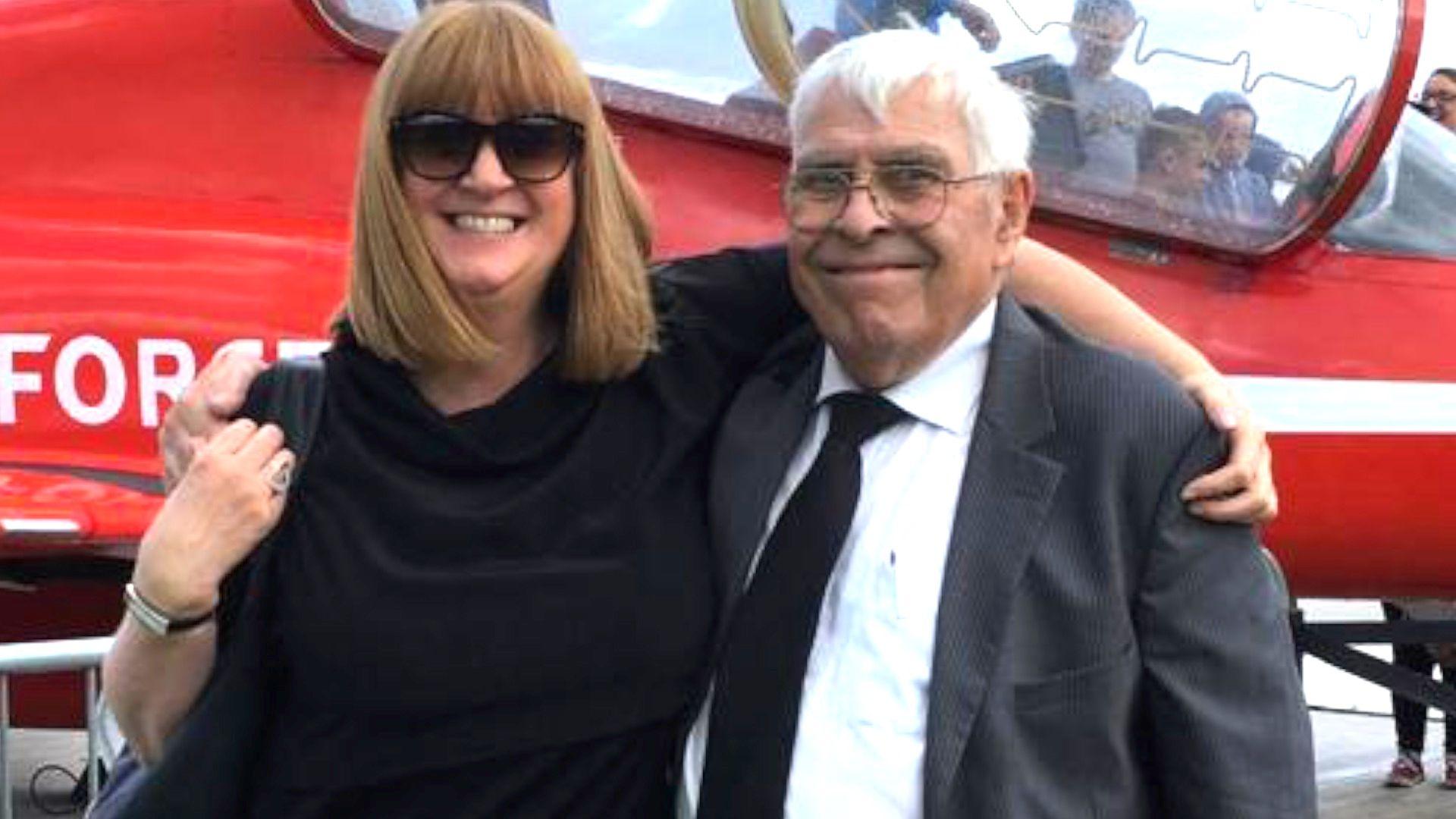 A picture of Melanie and Raphael Cull, melanie in black dress with sunglasses, raphael in black suit, black tie, white shirt and glasses, both smiling in front a red helicopter