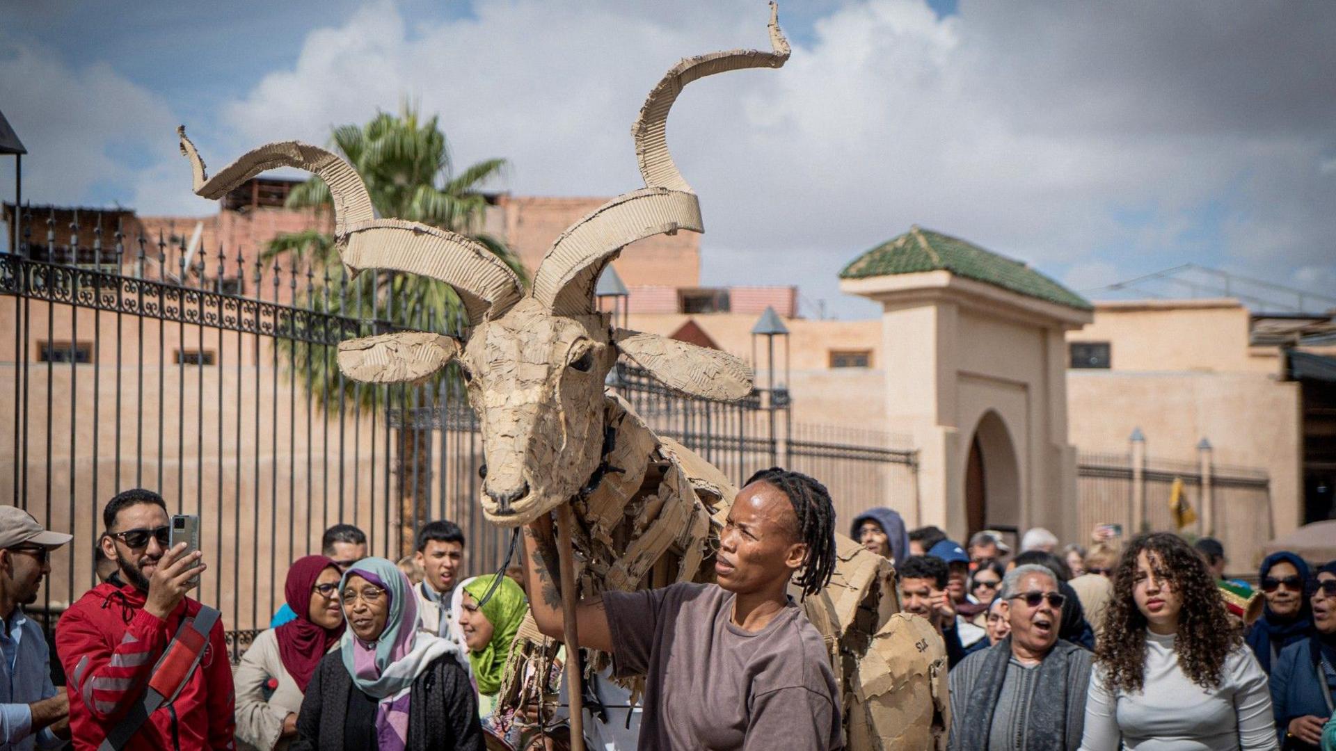 A large group of people following a large cardboard animal with winding horns in Marrakech.