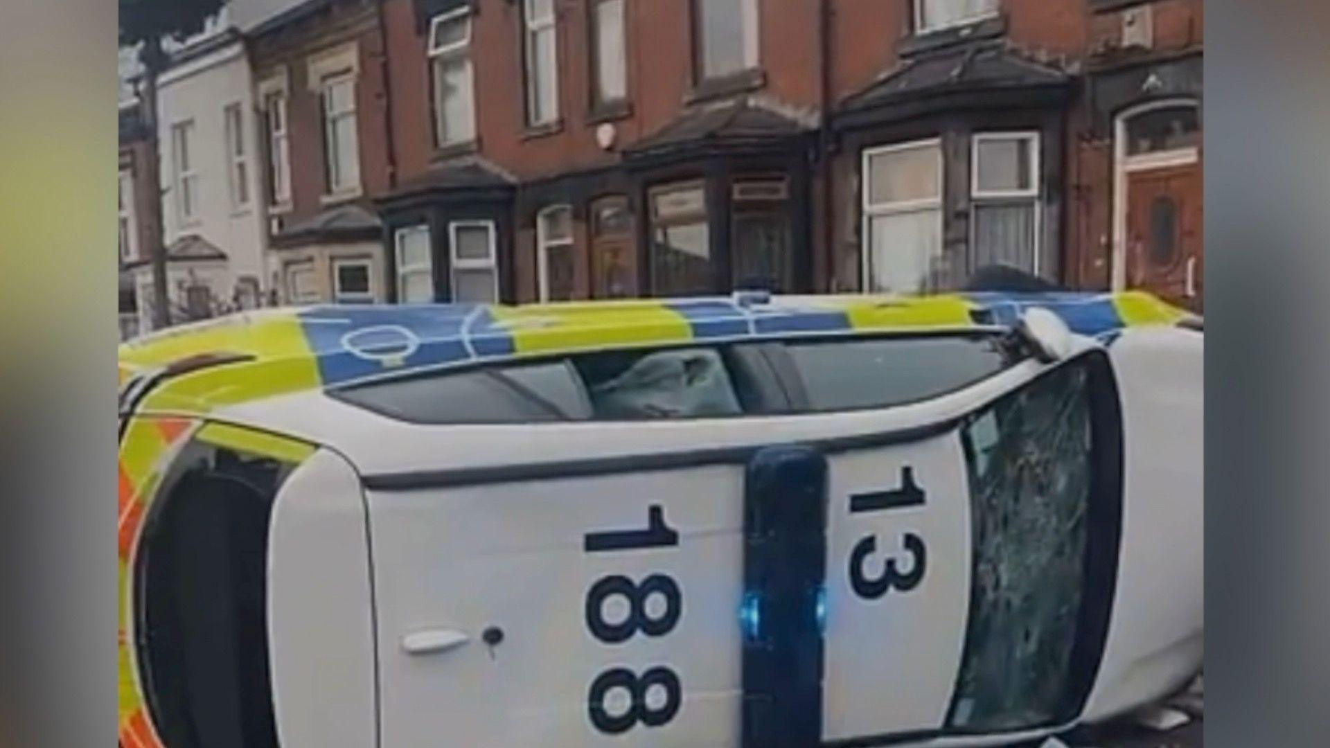 A police car with smashed windows lies on its side in the middle of a road.