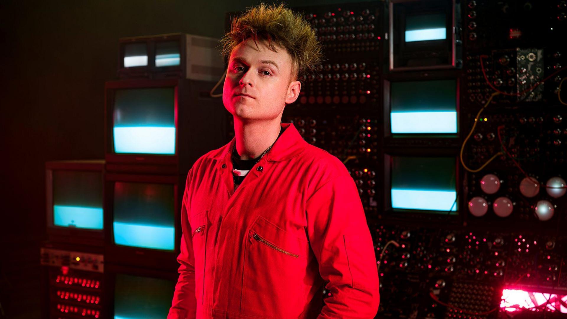 A white man has messy blonde/brown straight hair. He is stood in a big computer room with lots of wires and machines behind him. He is wearing a red boiler suit