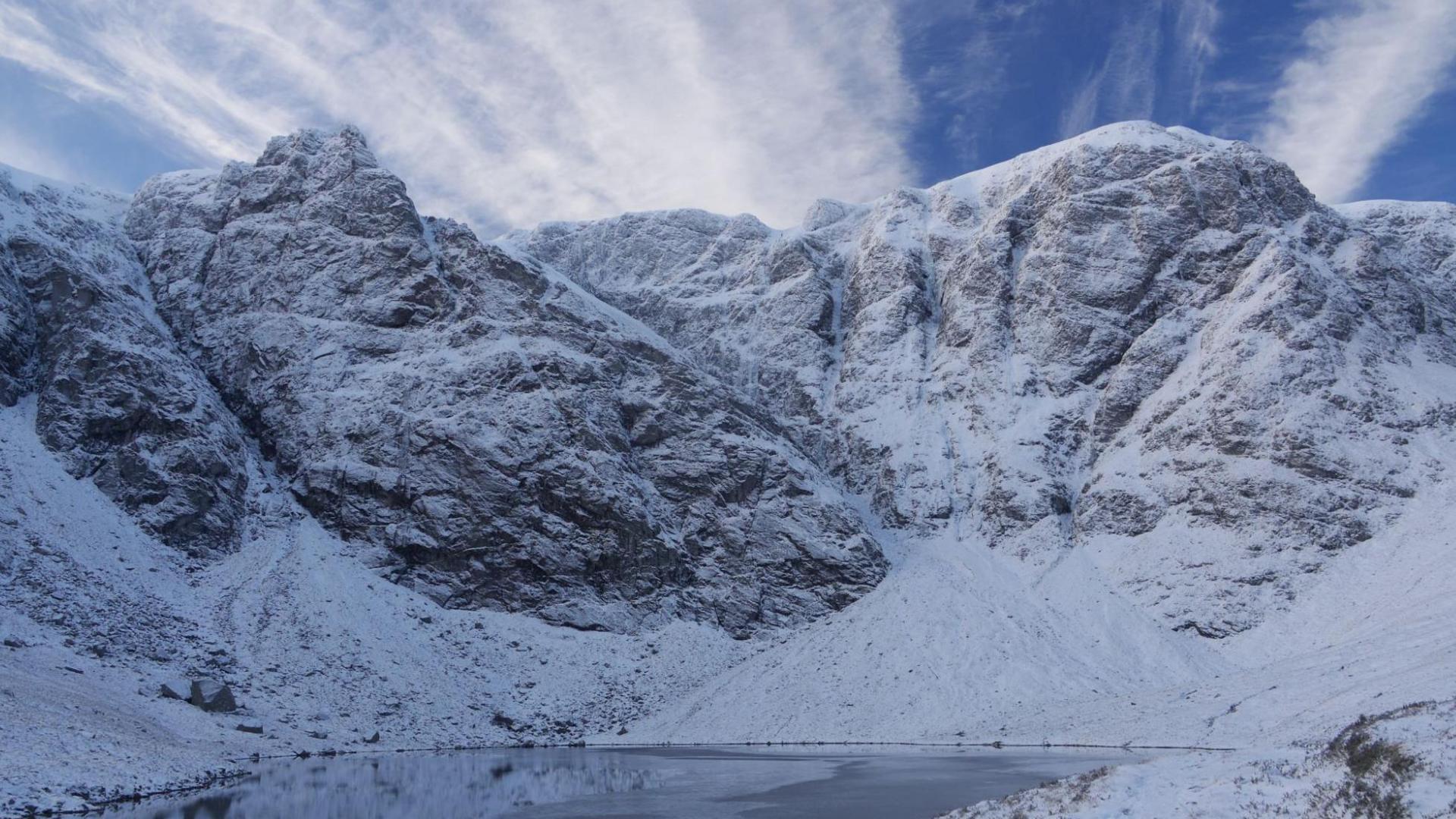 The craggy mountainside is dusted with snow. There is a lochan below the crags and wispy white cloud above the summits.