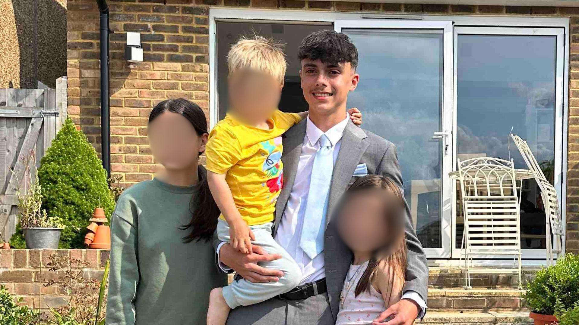 Brooklyn Bashford, wearing a suit and tie, hugs his younger sisters and lifts his younger brother.