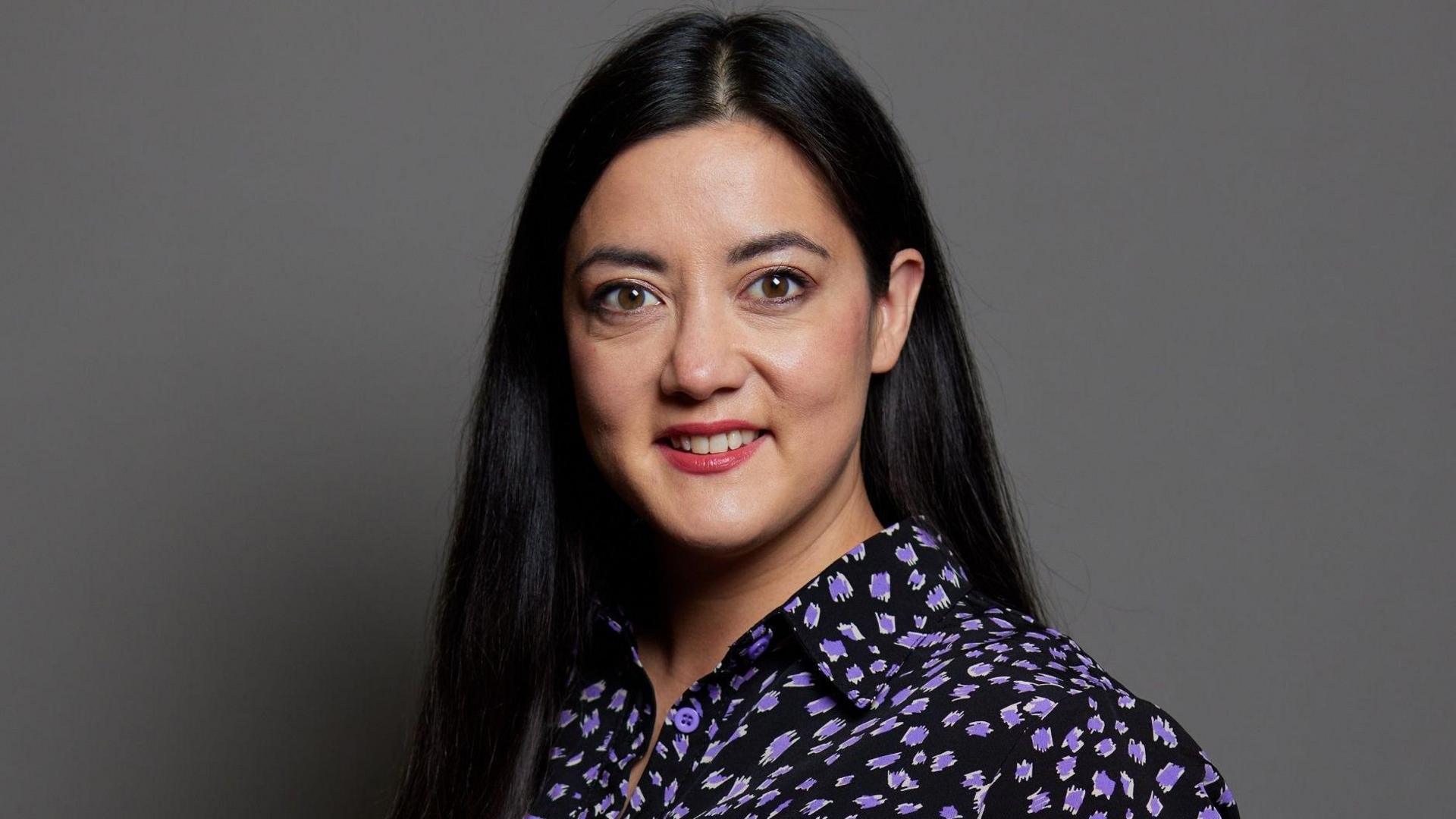 A portrait of Sarah Owen against a grey backdrop. She has black long straight hair, parted in the middle, and brown eyes. She is wearing a black shirt with purple shapes on it.