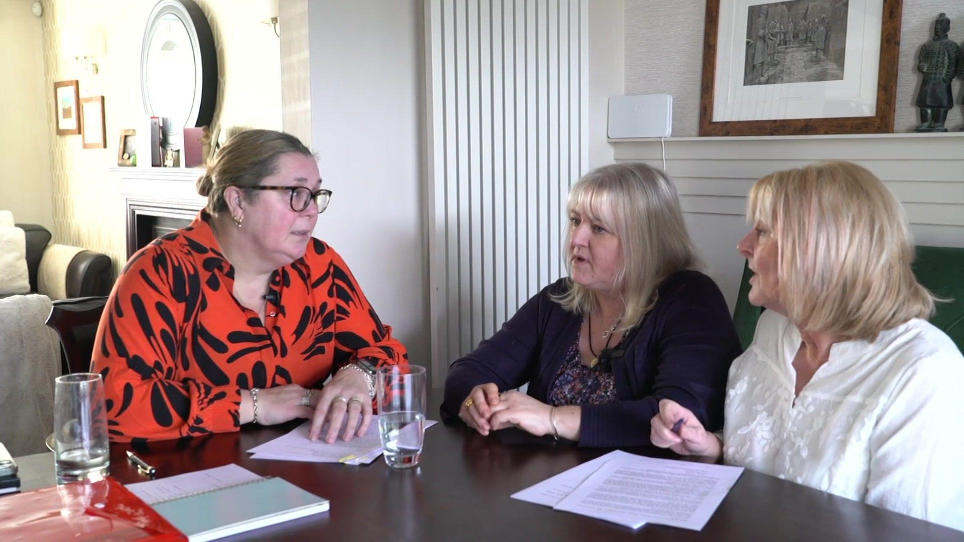 Barbara Cunningham, wearing a red and black blouse, Samantha Griffiths in a purple cardigan and Karen Paddock in a white blouse sit around a dark wooden dining room table chatting, with documents and glasses of water in front of them.