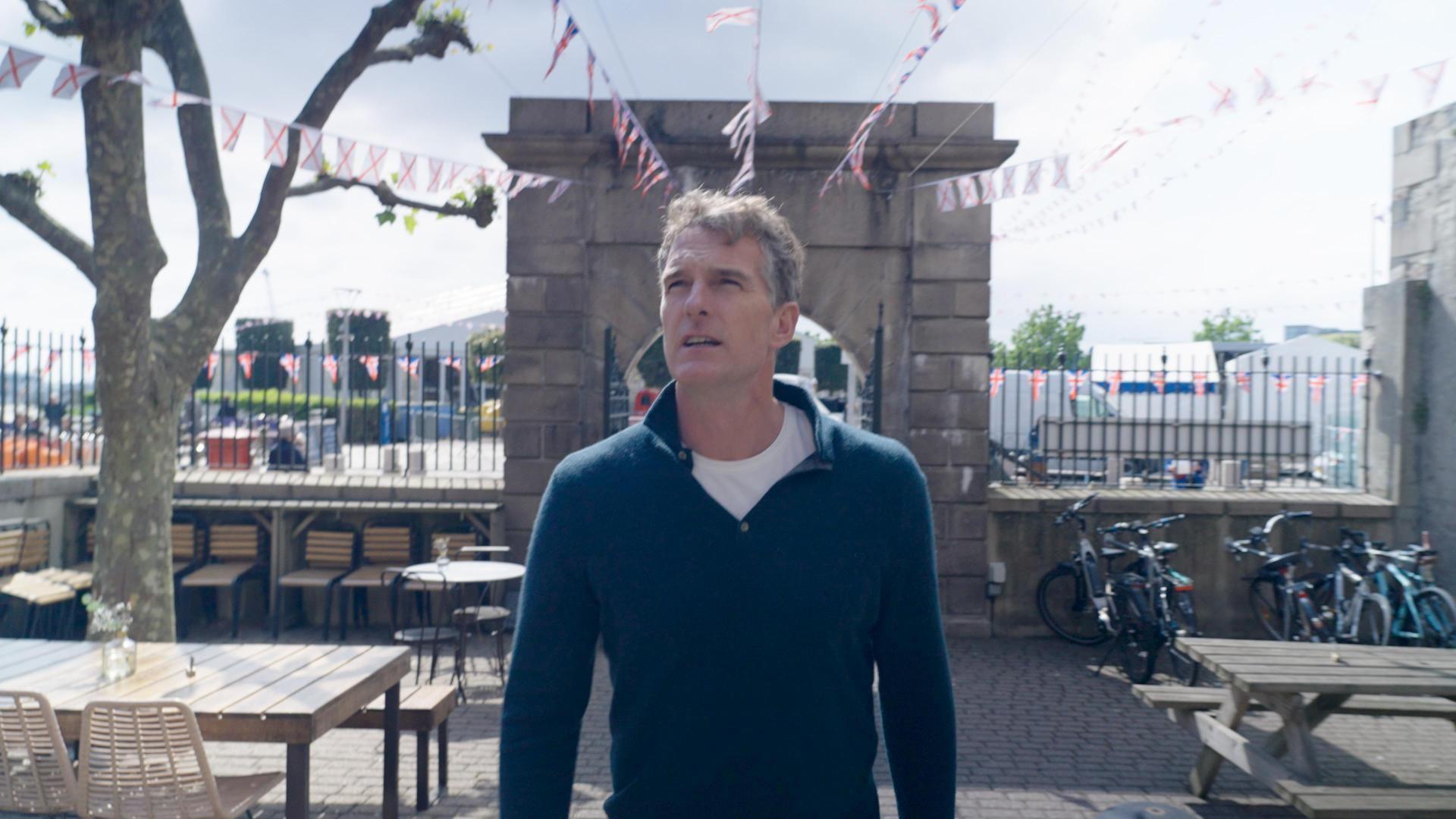Dan Snow is looking up as he stands outside Jersey Museum. He is surrounded by a stone wall, outdoor tables and Union Flag and Jersey flag bunting.