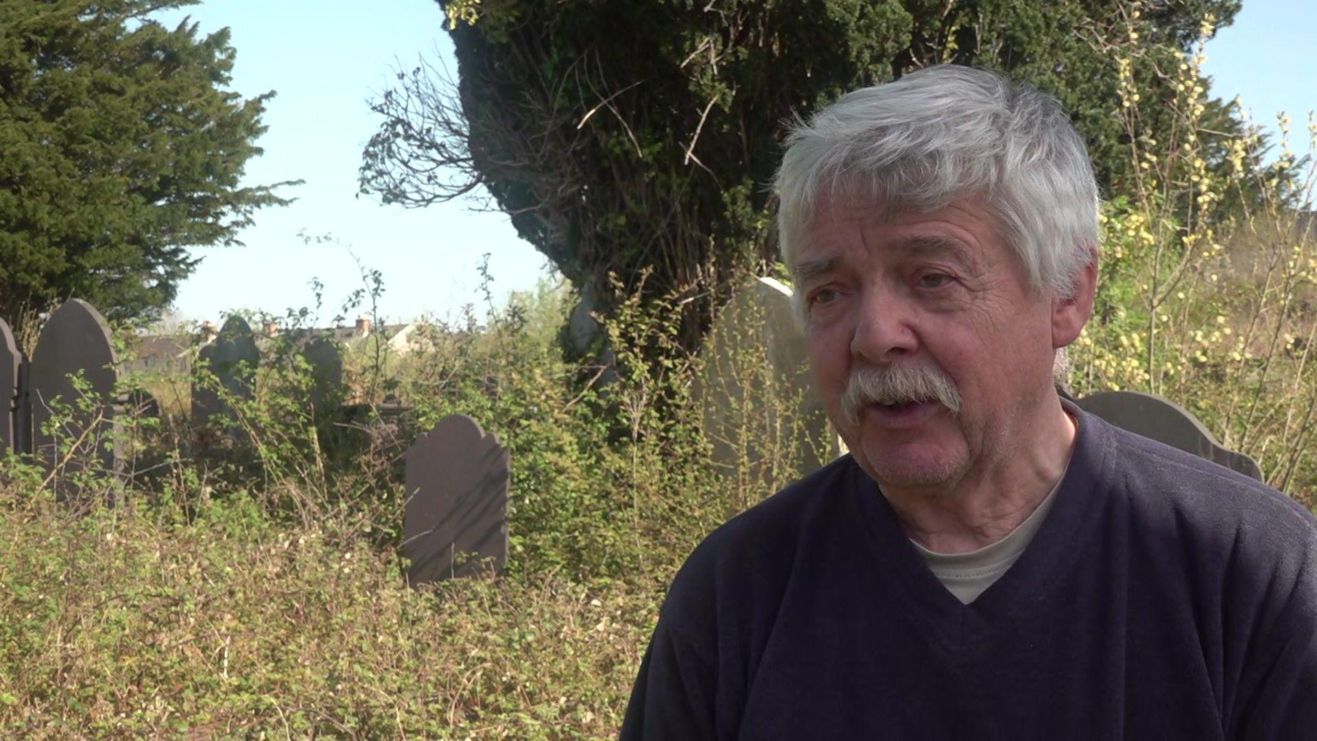 Gwyndaf Owen, dyn gyda gwallt a mwstash gwyn, yn cael ei holi ger cerrig beddi a thyfiant ar dir Eglwys Llanbeblig, Caernarfon