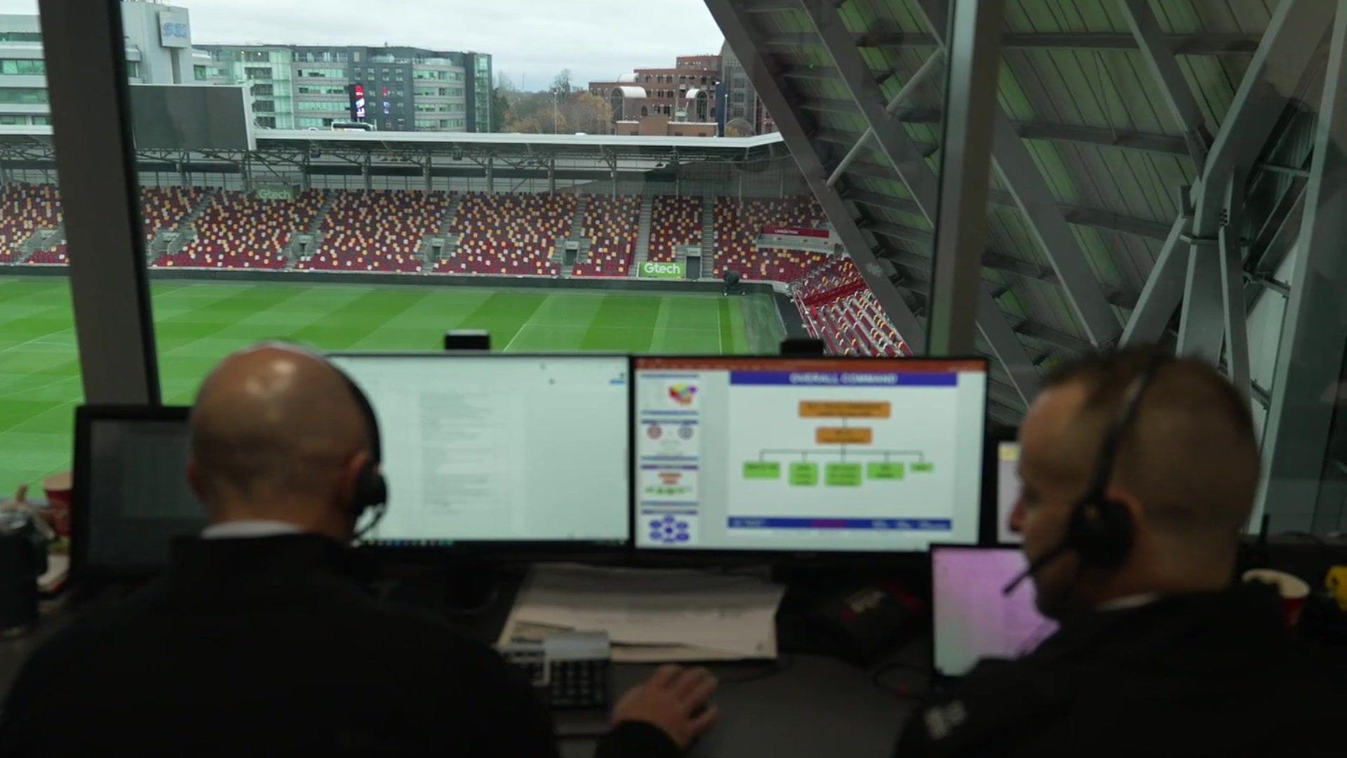  Two individuals wearing headsets sit at a command center overlooking an empty stadium, with multiple computer screens displaying  operational information.