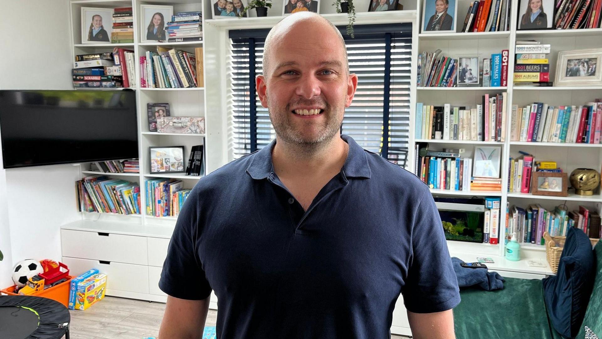 Stef smiles at the camera, standing in the living room with the bookshelves behind him. There are several school photos of his daughters on the shelves above. He wears a blue open necked collared airtex-style shirt.
