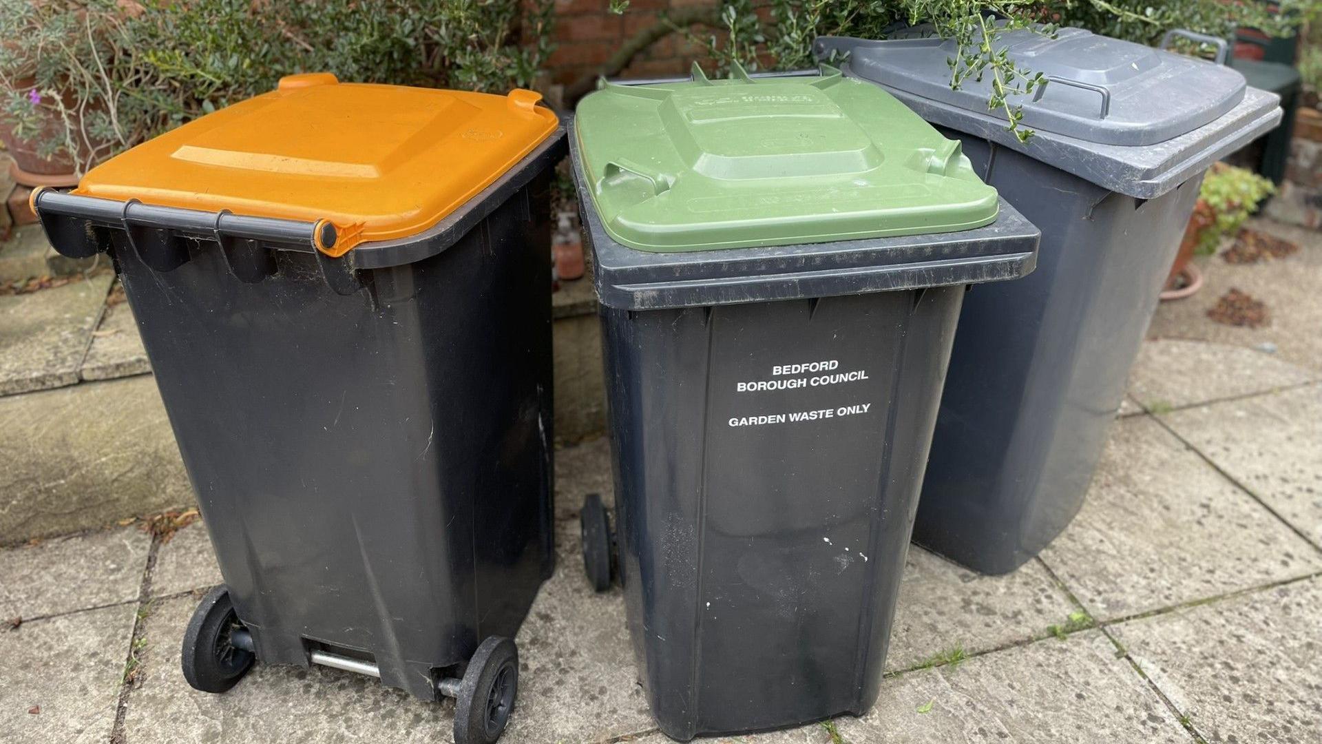 Three grey wheelie bins - with orange, green and grey lids. The green-lidded bin says "Garden Waste Only" in white lettering. The bins are standing on grey paving slabs with a hedge behind.