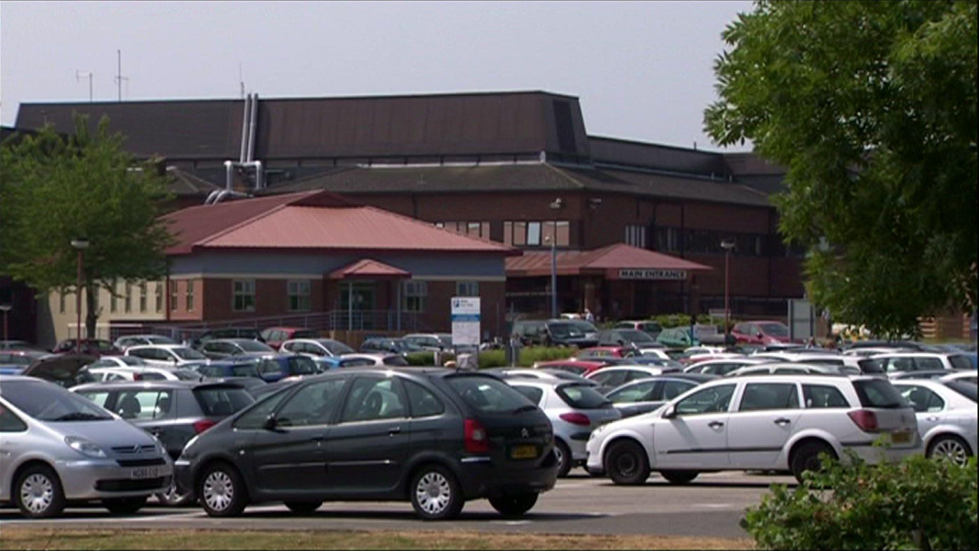 Cars parked in car park with a dark brown hospital building in the background.