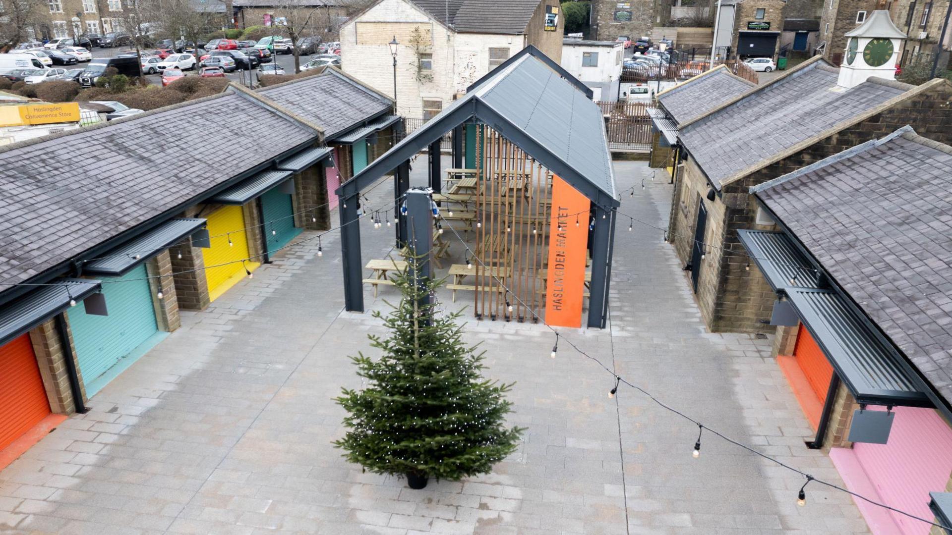 A drone shot of the inside of the market. There are brightly coloured shacks flanking a shack in the middle of the grounds. There are Christmas lights hung between each building and a Christmas tree in the middle.