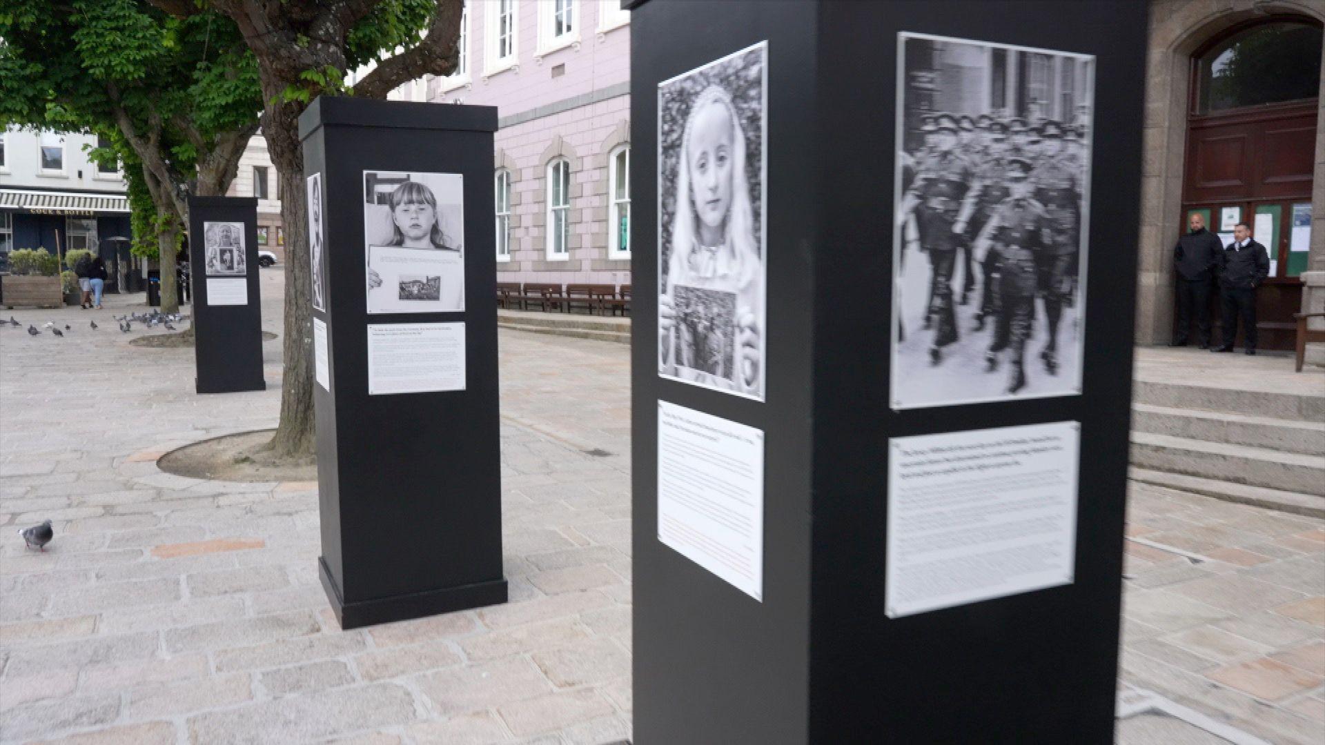 A picture of plinths in Jersey's royal square. They are black with black and white images on. They are surrounded by buildings and trees.