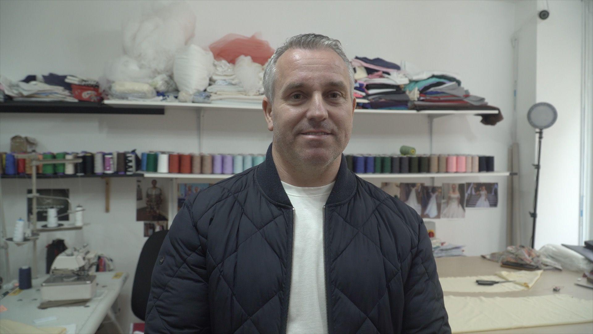 A man with greying hair is wearing a black puffer jacket and a white shirt. He is standing in front of a working table ladened with sewing equipment including a sewing machine, scissors, and sewing thread rolls.
