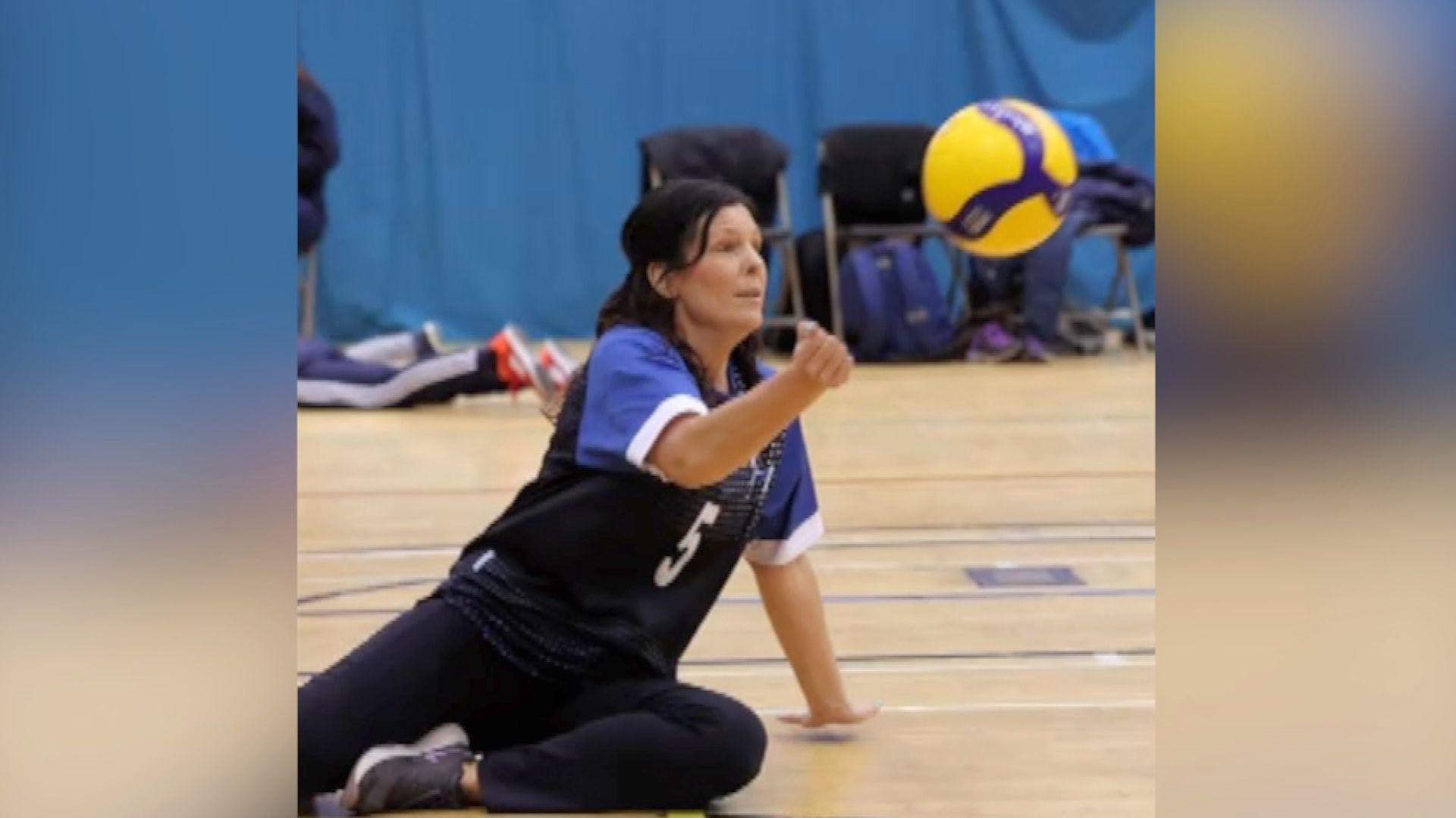 Kerry Snuggs is in a sports hall. She is sat on the floor, wearing a blue and black sports jersey and black sweat pants and trainers as a yellow and purple volleyball flies towards her. Her right hand is raised as she prepares to hit the ball.