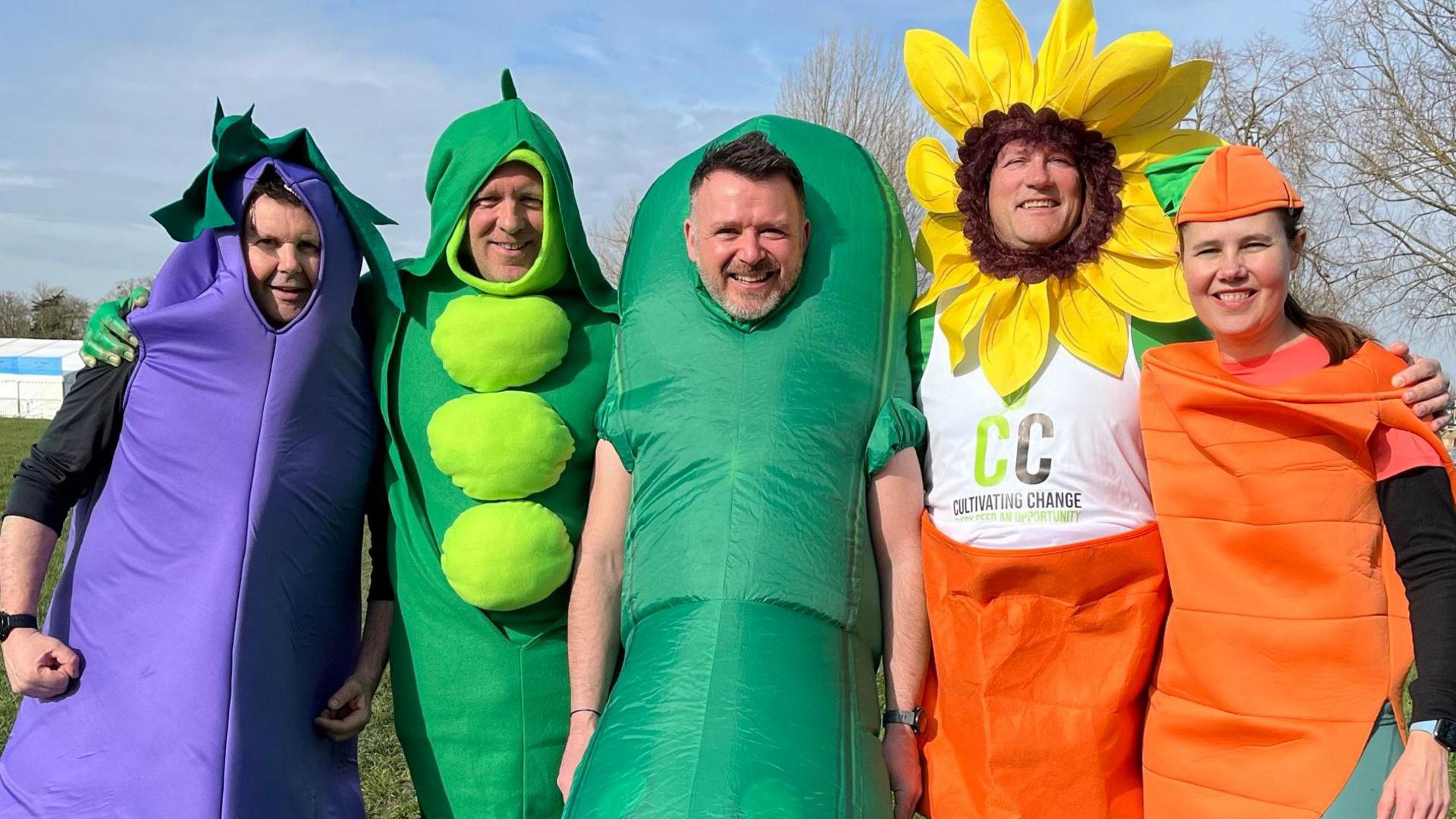 A group of four men and a women dressed as various vegetables and one as a sunflower. They smile at the camera.