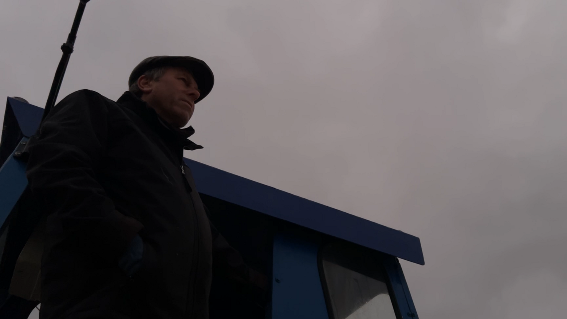 Alan Stead standing against a grey and cloudy sky looking out to sea. The cab of a blue tractor is visible behind him.