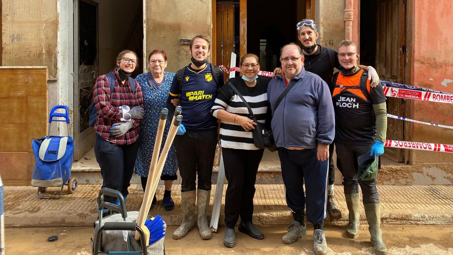 A group of volunteers wearing wellies and gloves, standing in front of a building with three open doors in Valencia. They are posing with their arms around each other and smiling at the camera. They are there to help with the clean up efforts. In front of them there is a bucket full of brooms and cleaning supplies.