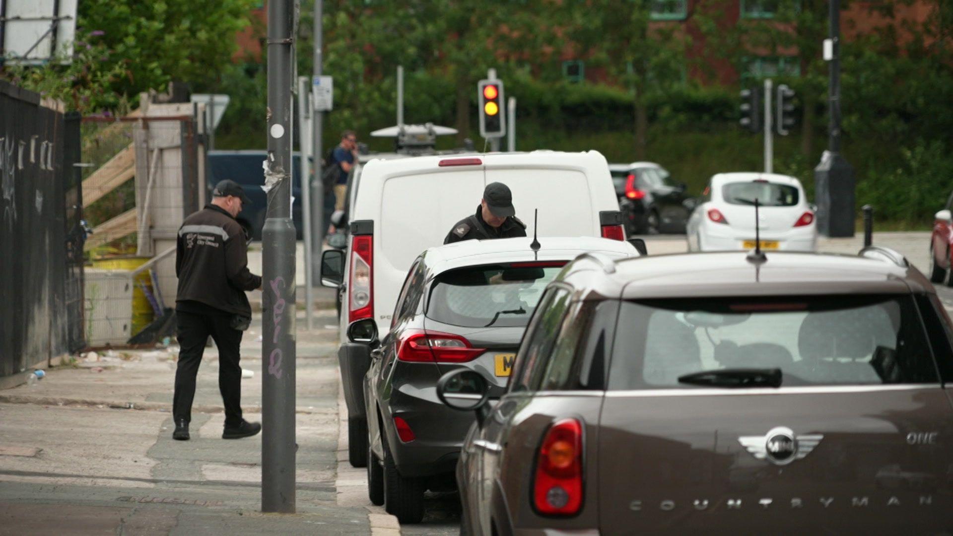 Two traffic wardens operating in Liverpool's Baltic Triangle area, checking cars and vans parked along the road.