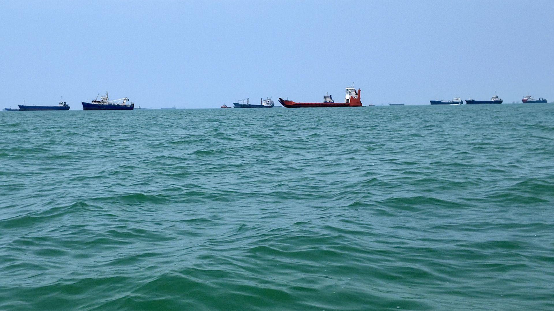 At least 10 large ships out at sea. The water is a bit choppy and the sky is blue.