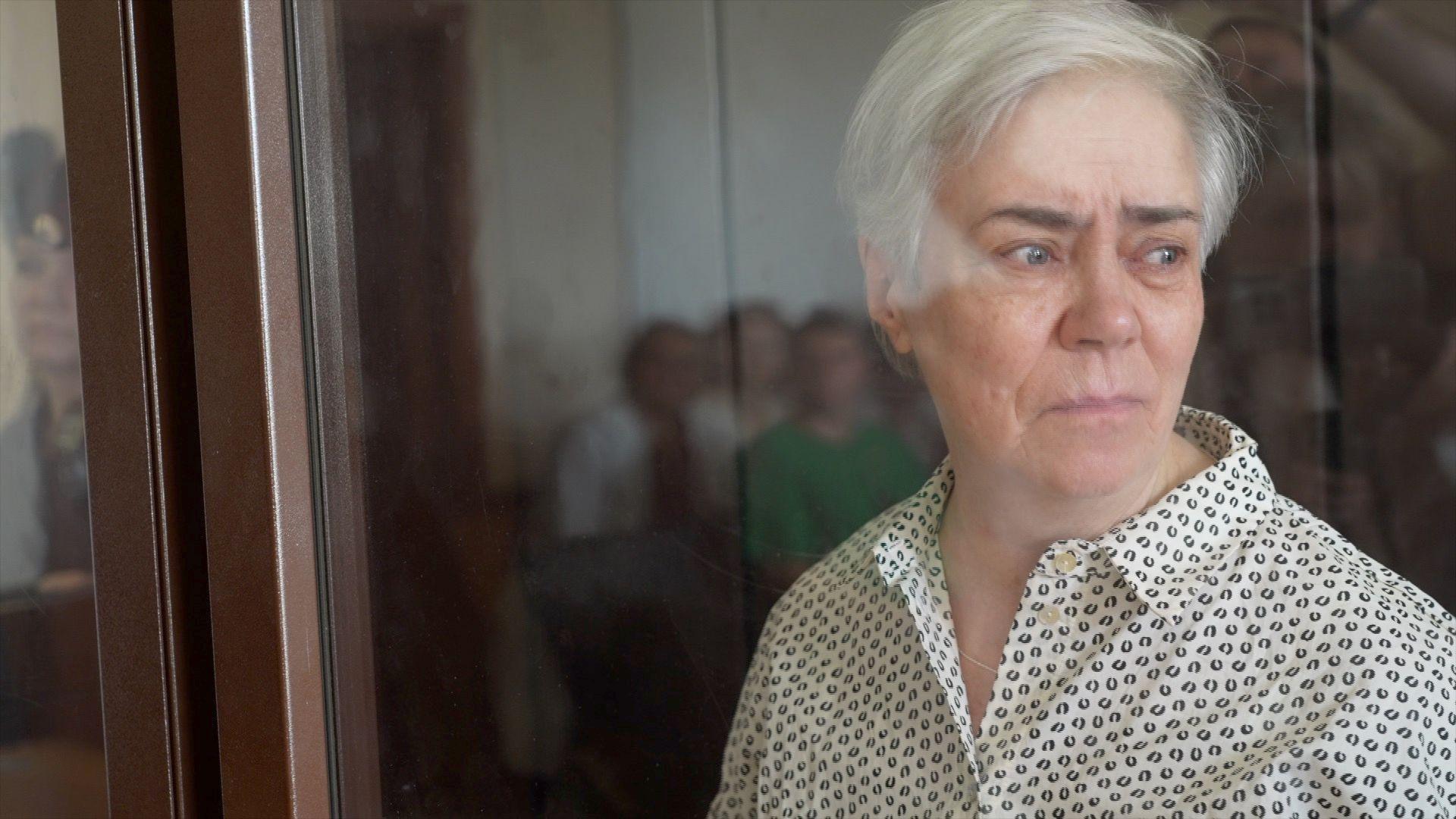 Nadezhda Buyanova, who is also pictured behind the glass of a court dock, looks to the right with a serious expression on her face. She has short grey hair and dark eyebrows, wearing a white shirt with a black dotted pattern.