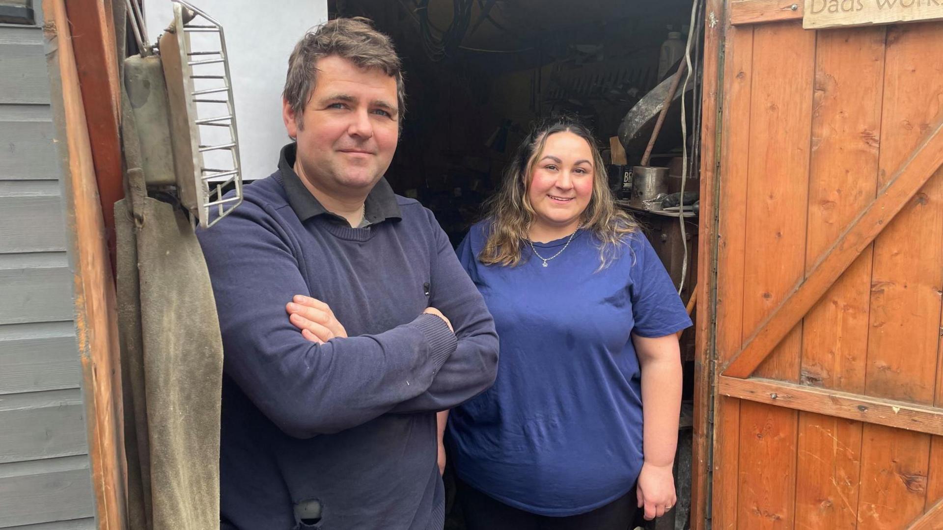 A young man and woman stand smiling in the doorway of a garden shed