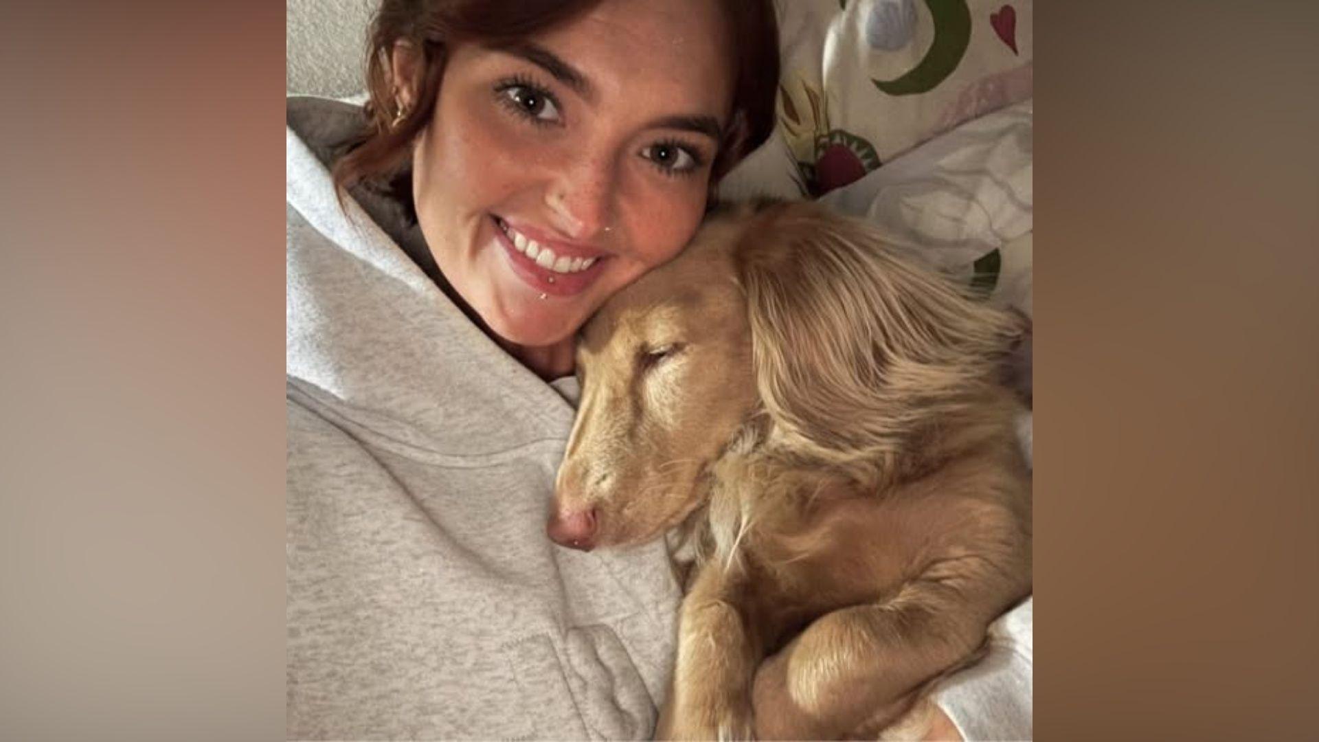 Jess McKee wearing a grey tracksuit cuddles a sandy-coloured dog. She is smiling. 