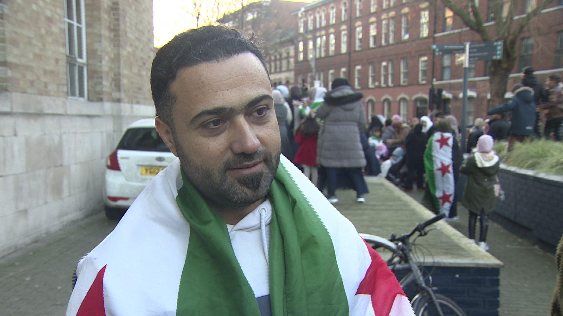 Mazen Haseno draped in a Syrian flag, stood outside broadcasting house with a crowd of people visible in background celebrating