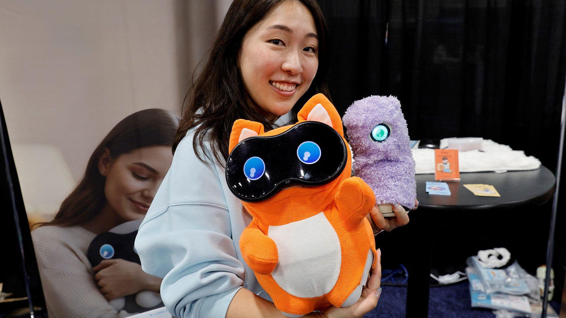 A person stands holding two tech toys, which look like stuffed animals with digital eyes 