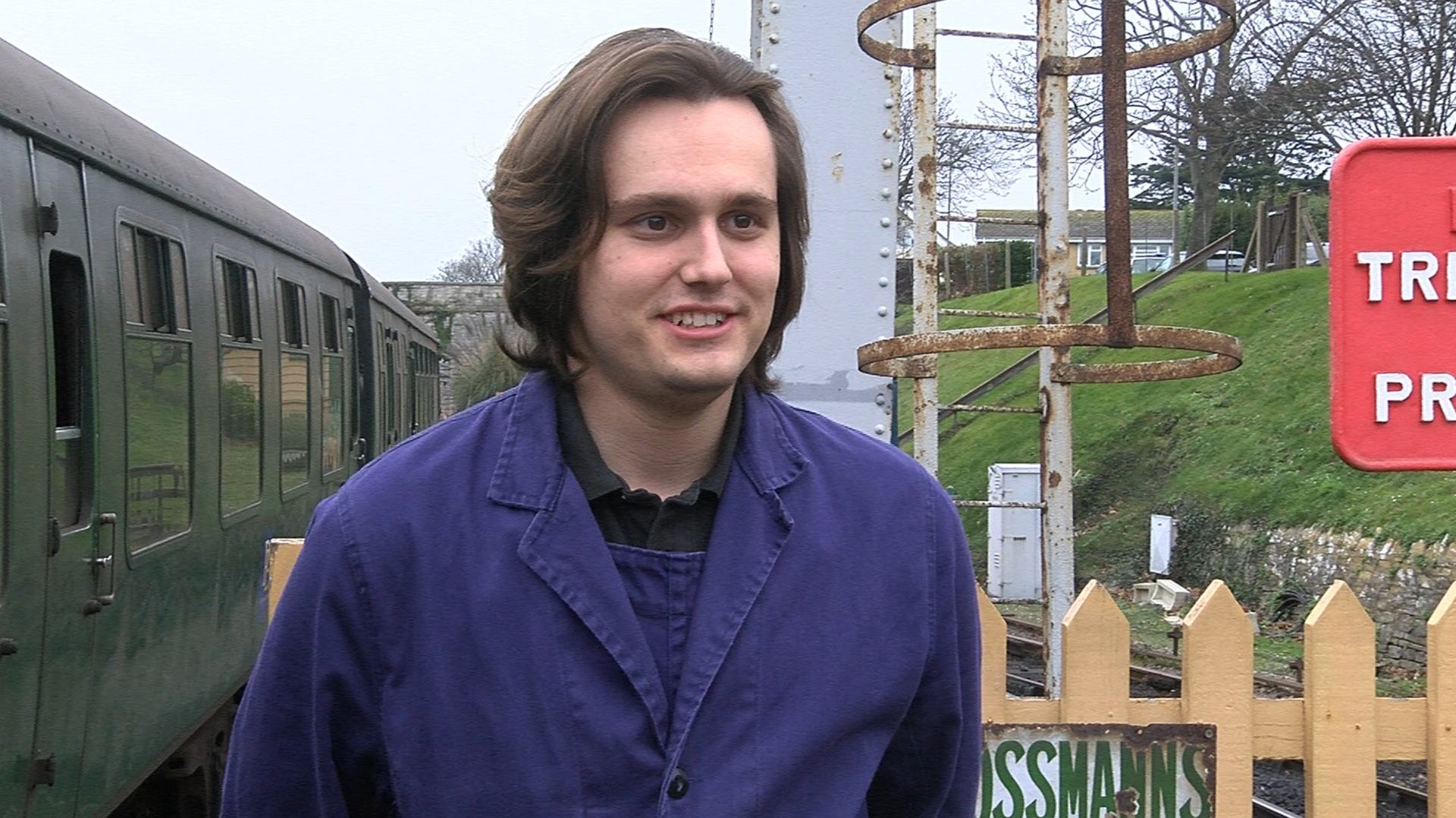 A young man with dark hair wearing dark blue overalls with a green train carriage and a railway behind him.