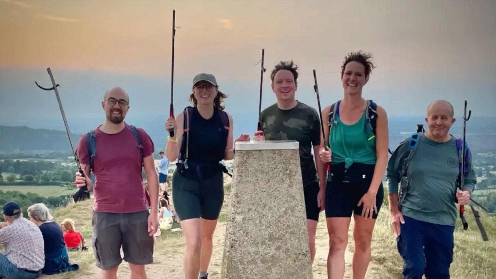 People stood at the top of a hill in Gloucestershire with litter pickers in hand. They are smiling at the camera and the picture is taken just after dawn
