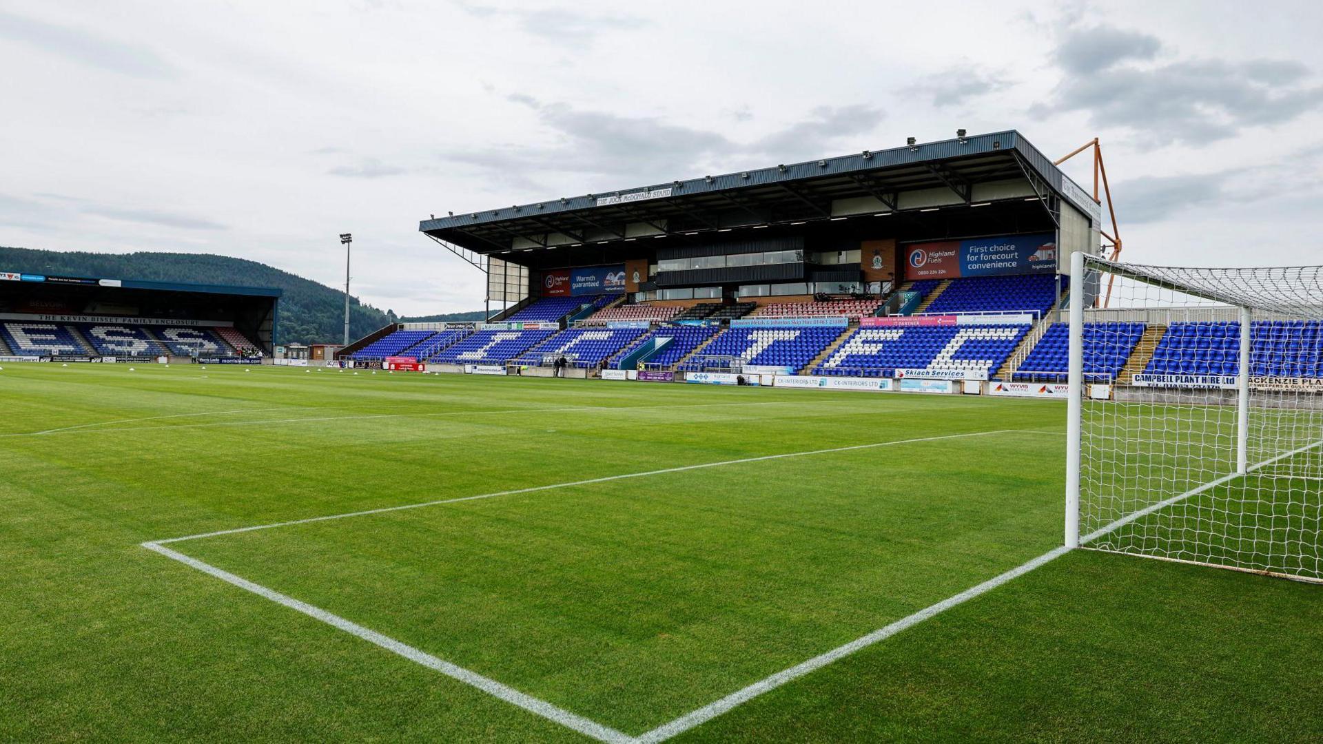 The exterior of the stadium's main stand, a large structure with a sloping metal roof. Rows of blue plastic seats along the front of the stand have the initials ICTFC in white. In the foreground is the pitch and a set of goals.