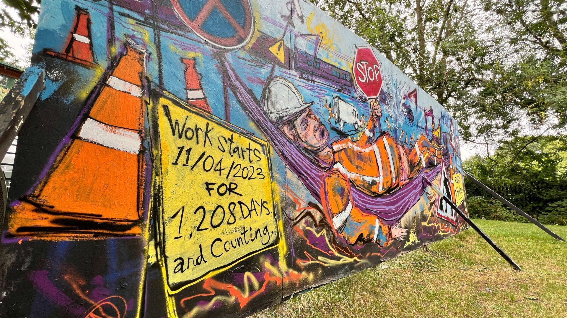 An image of the mural, which is on a large board propped up by long stick in the middle of a patch of grass, with some trees and hedging in the background. The mural shows a caricature of a workman wearing an orange high-vix long sleeved jacket and trousers, with a white helmet. He is lounging on a purple fabric hammock and holding a red and white 'STOP' sign. In the background, which is painted blue, there are some red flags and orange and yellow traffic cones. In the foreground of the image there is a yellow road sign which reads 'Work starts 11/04/2023 for 1,208 days and counting.'