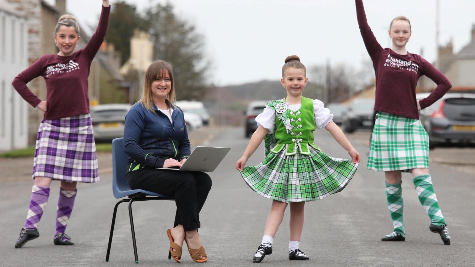 Dance teacher Tanya Horne has shoulder length fair hair and is sitting on a chair outside. She has a laptop on her lap and is surrounded by three of her Highland dancers. The girls are wearing different coloured tartans.
