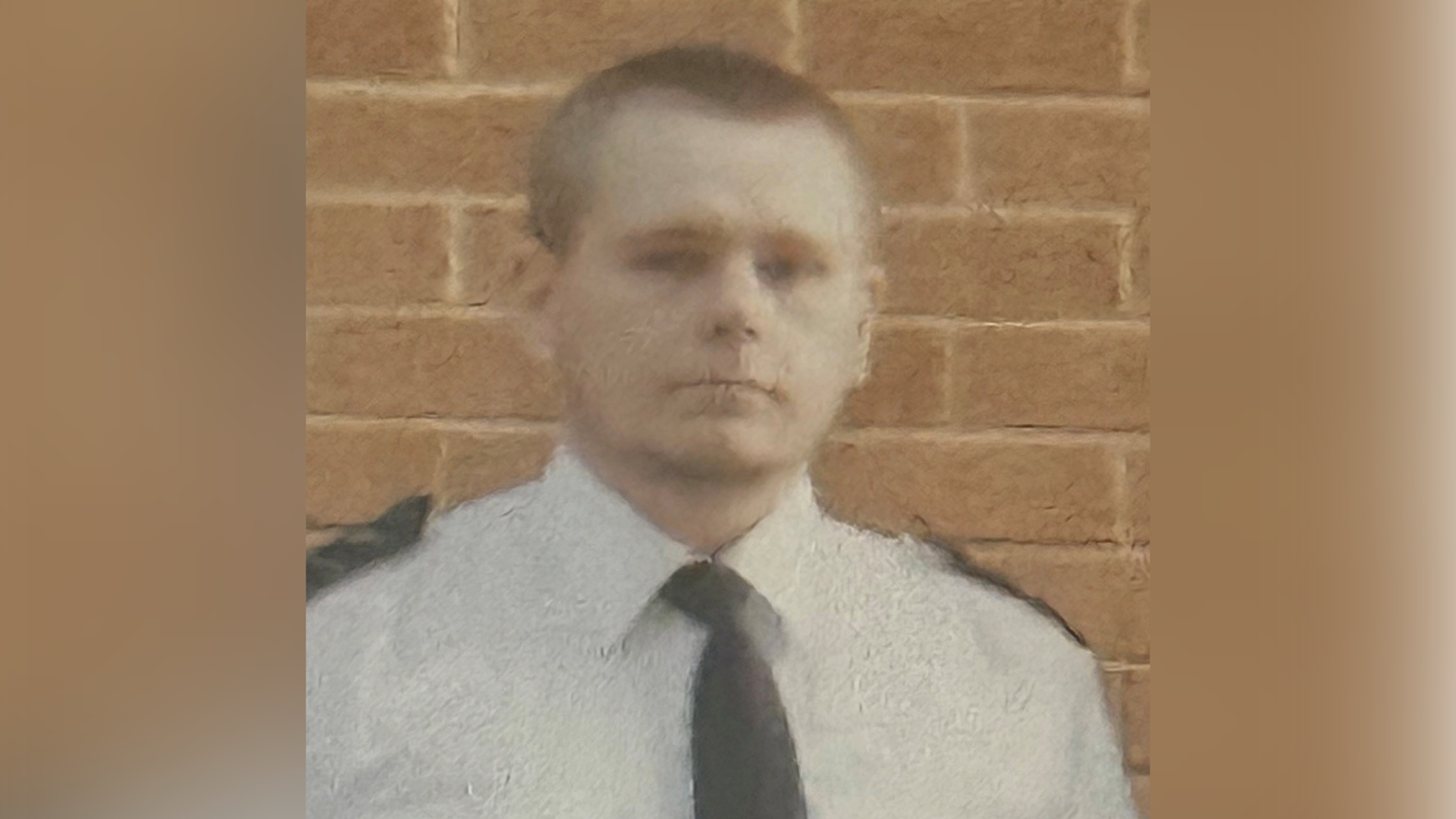 A man with short brown hair standing in front of a brick wall. He is wearing a white shirt, black tie, and there are black patches on his shoulders