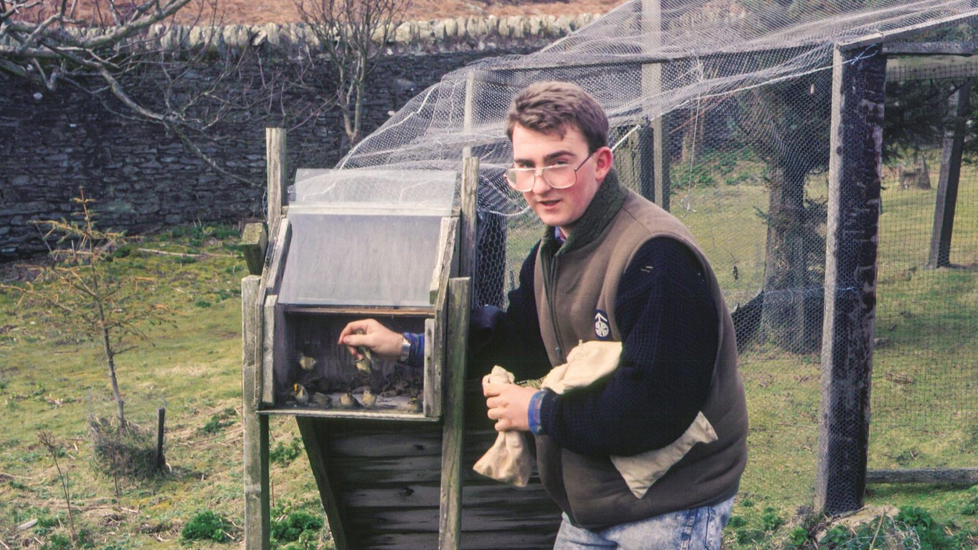 A photo from the 80s of Aron, at the time a young man who is wearing large glasses and a camouflage gilet and jeans as he handles a bird. There is a large net in the background.