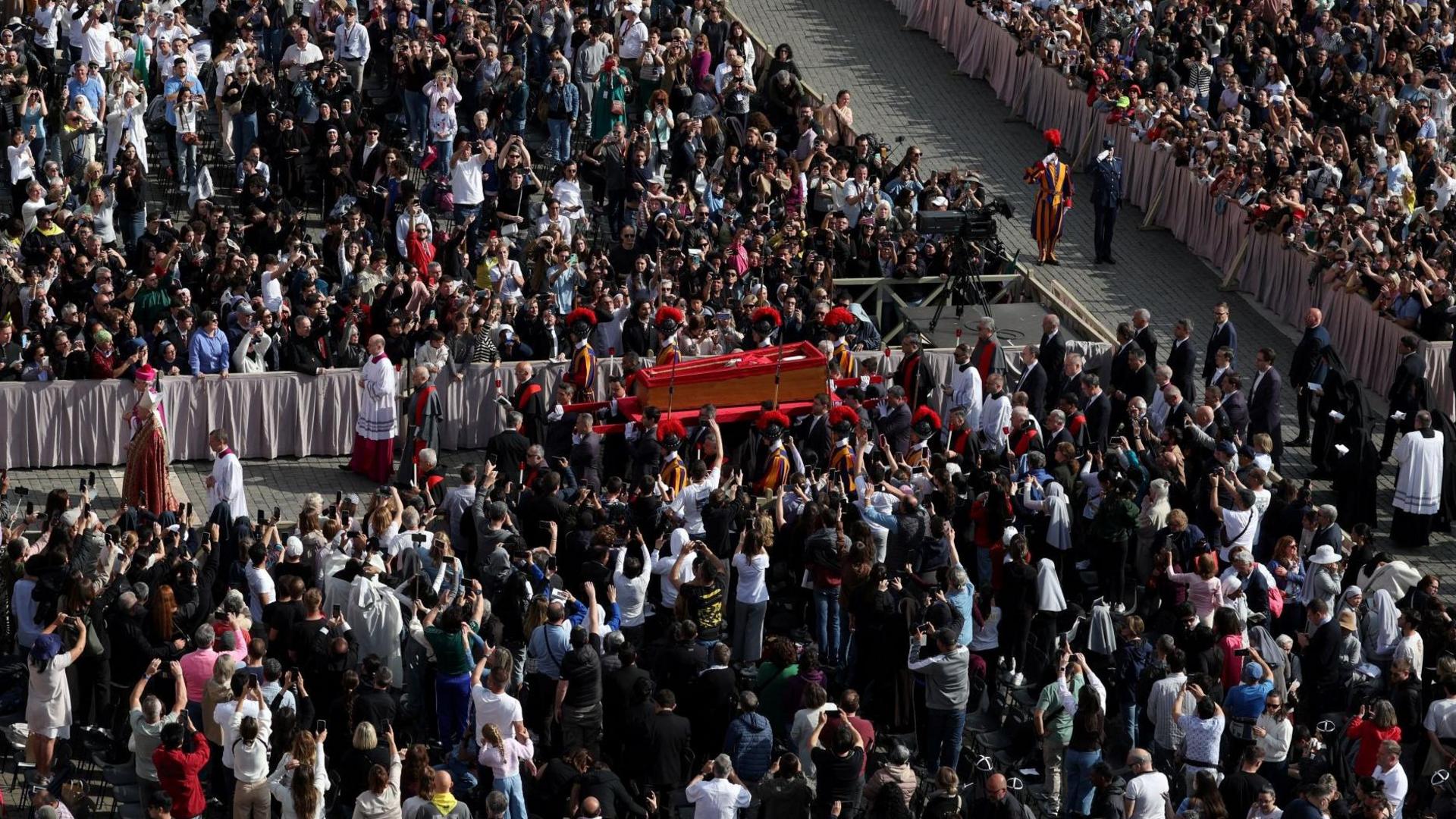 A wide shot of the Pope's open coffin proceeding through St Peter's Square. There are thousands of people lined around the processional route as clergymen and guards carry the coffin of Pope Francis.