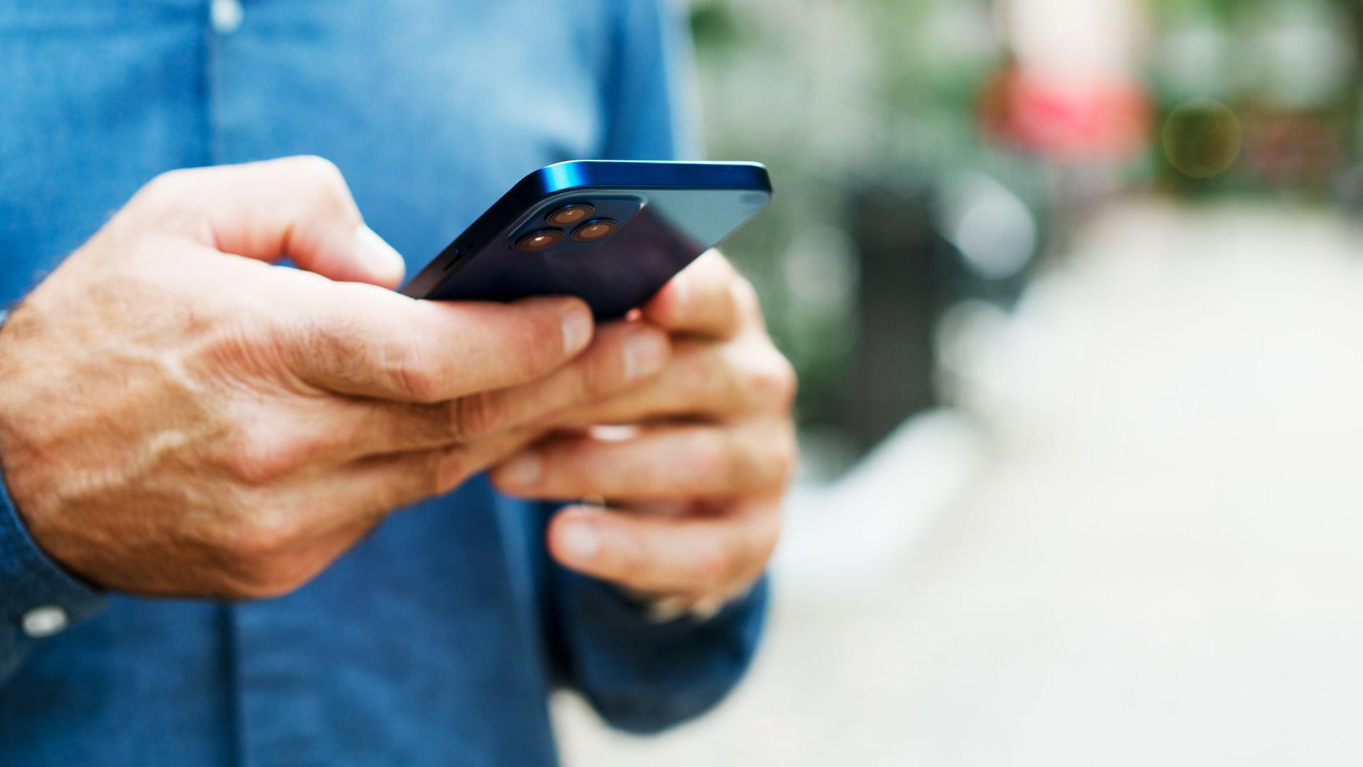 A man using a mobile phone in the street. He is wearing a blue shirt, with the background of the image blurred.