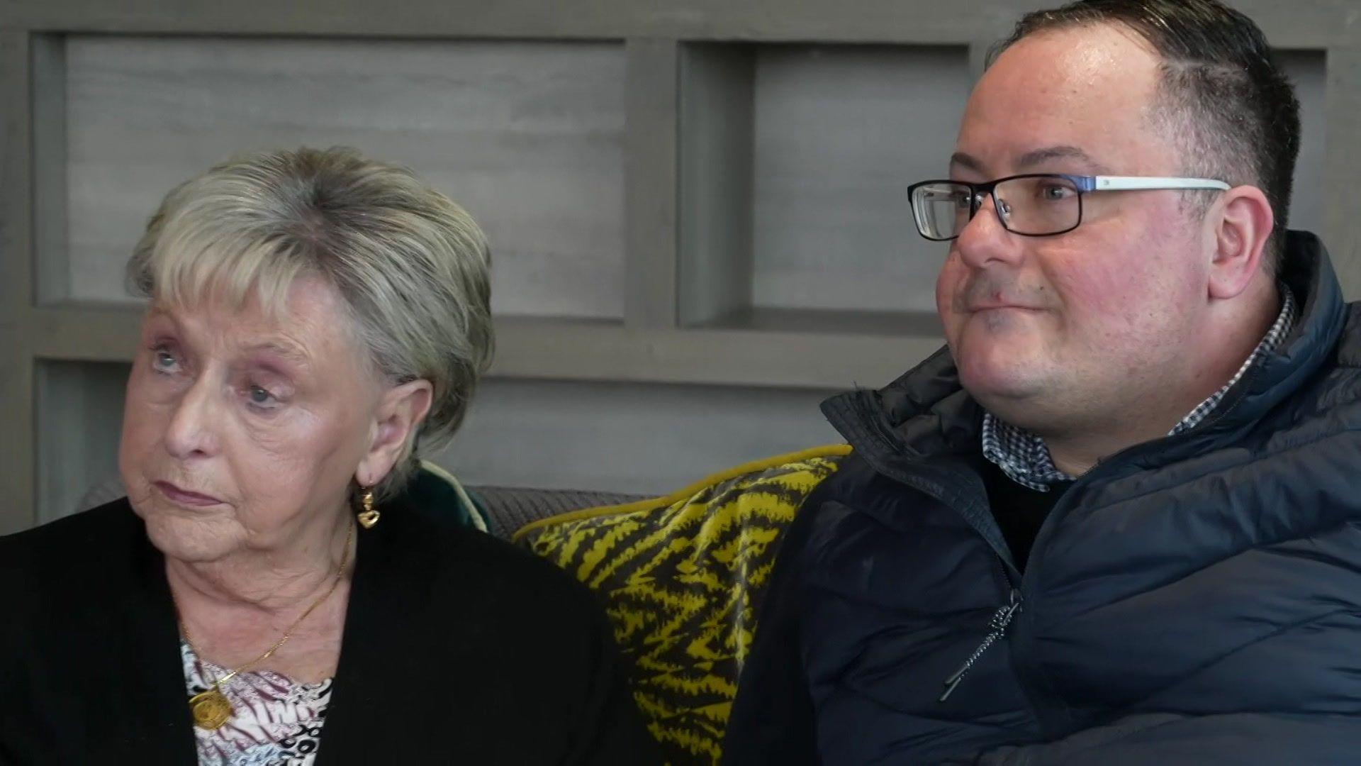 Ann and Kevin sit side by side on a sofa. She has short blond hair and is wearing a black jacket over a white floral shirt. He has short dark hair, is clean shaven and is wearing glasses, a blue and white check shirt, black jumper and navy padded coat.