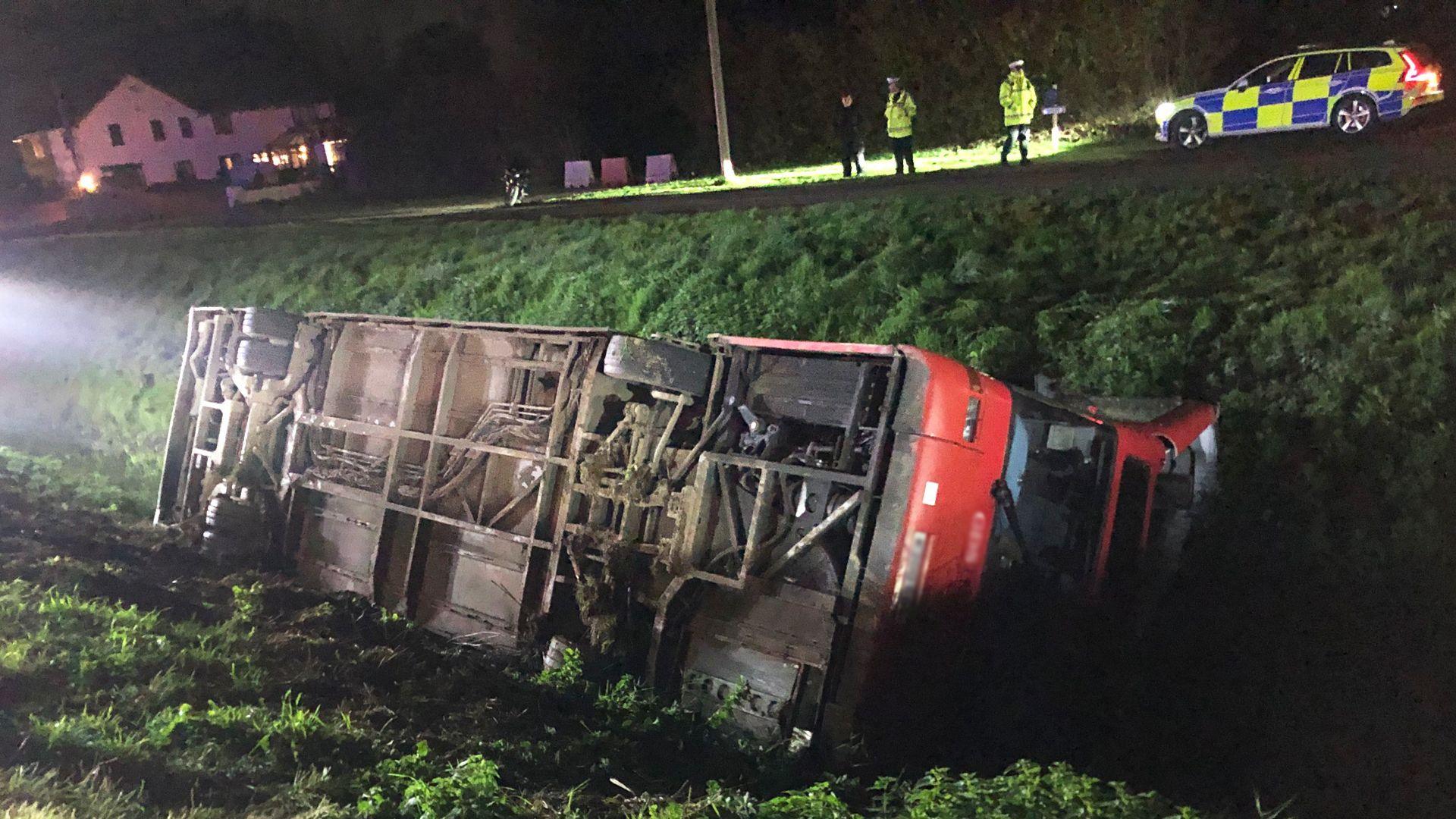 A red double-decker bus in a ditch at night-time. It is on its side and its roof has been crushed in. It is being lit up by emergency service's lights. In the background on the opposite bank are police officers and a police car.