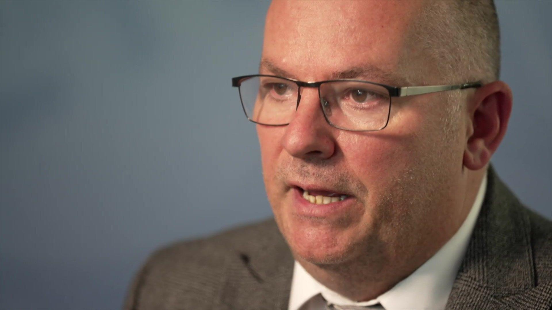 Det Sgt Simon France speaking to the camera; he is bald, wearing glasses and a grey suit.