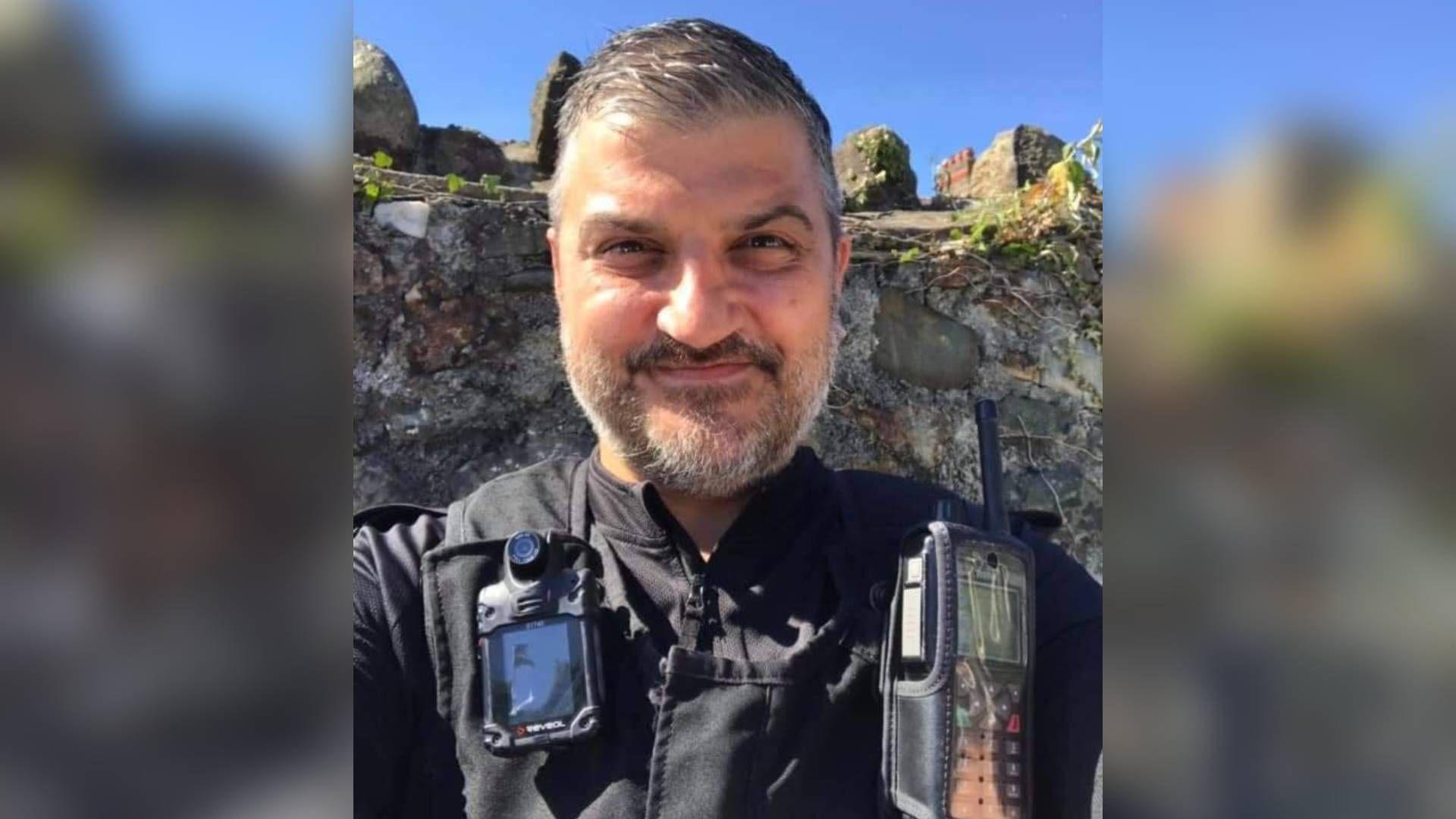 Picture of former policer officer Eurwyn Thomas in his uniform. He smiles towards the camera, a bright blue sky can be seen in the background behind a grey stone wall. He is of south-asian ethnicity, has brown eyes and a brown hair and beard speckled with silver hairs. 