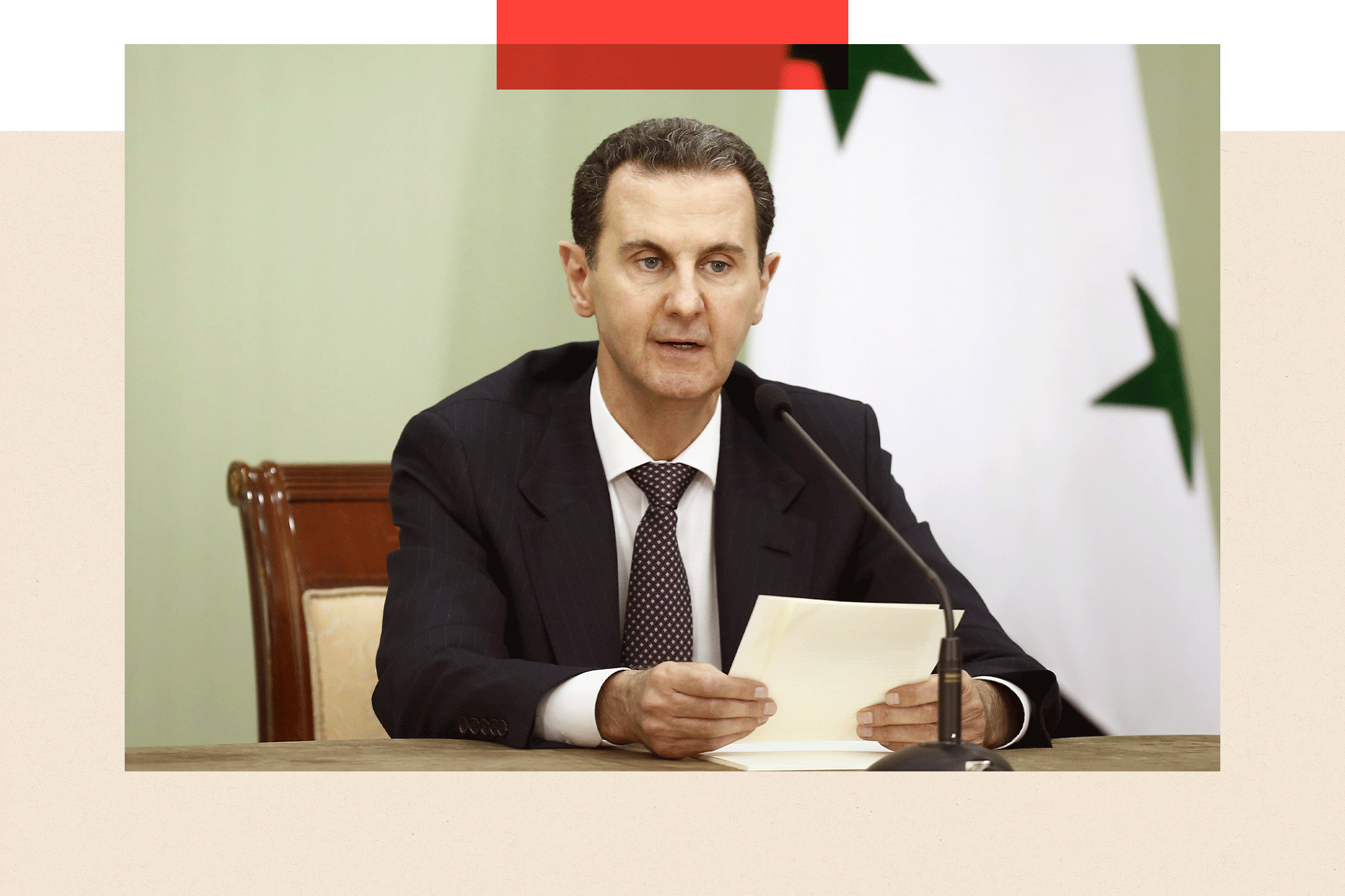 Bashar al-Assad sits behind a microphone with papers in his hands, with a flag behind