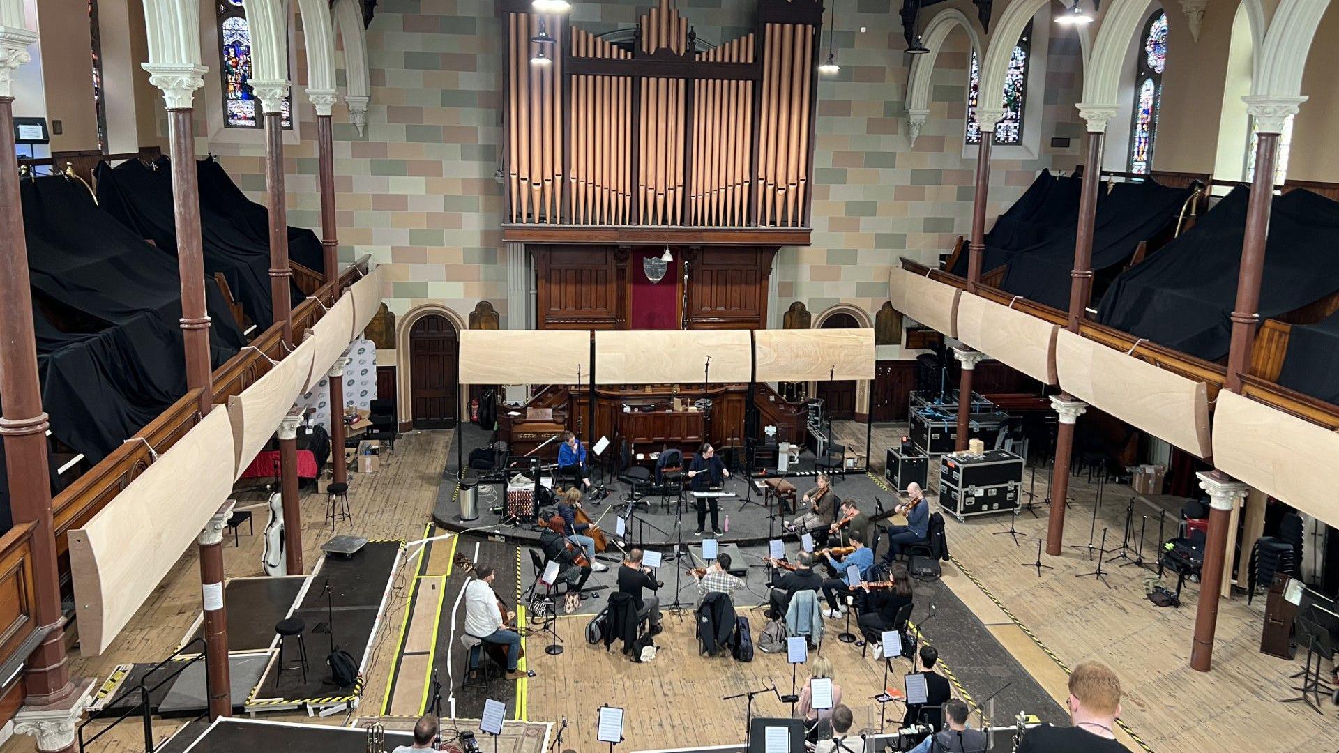 Orchestra rehearsal down on the ground floor of a church. Picture is taken from a balcony, which shows the entirety of the church with stain glass windows. 