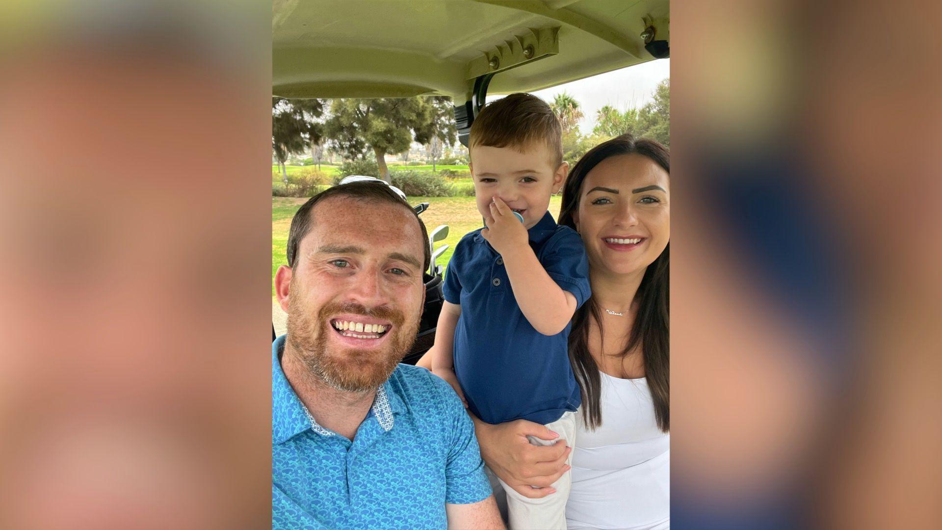 A family photo shows dad Luke on the left, Wilson in the middle and Rebecca on the right. They are all looking at the camera and smiling, while sat in a golf buggy on a golf course. Luke has short hair with a ginger beard and wears a bright blue printed polo shirt. Wilson is standing, holding a blue dummy to his mouth and has short light brown hair. He wears a navy blue polo shirt and white trousers. Rebecca has her arm around Wilson's waist, she has long brown hair and wears a round neck white t shirt.