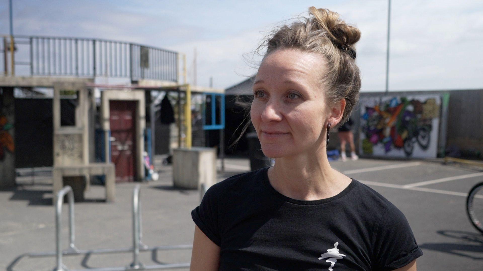 Anja Meinhardt wears a black top and looks slightly ahead of the camera. She has a smile on her face and her hair is up in a bun. She is standing in a playground